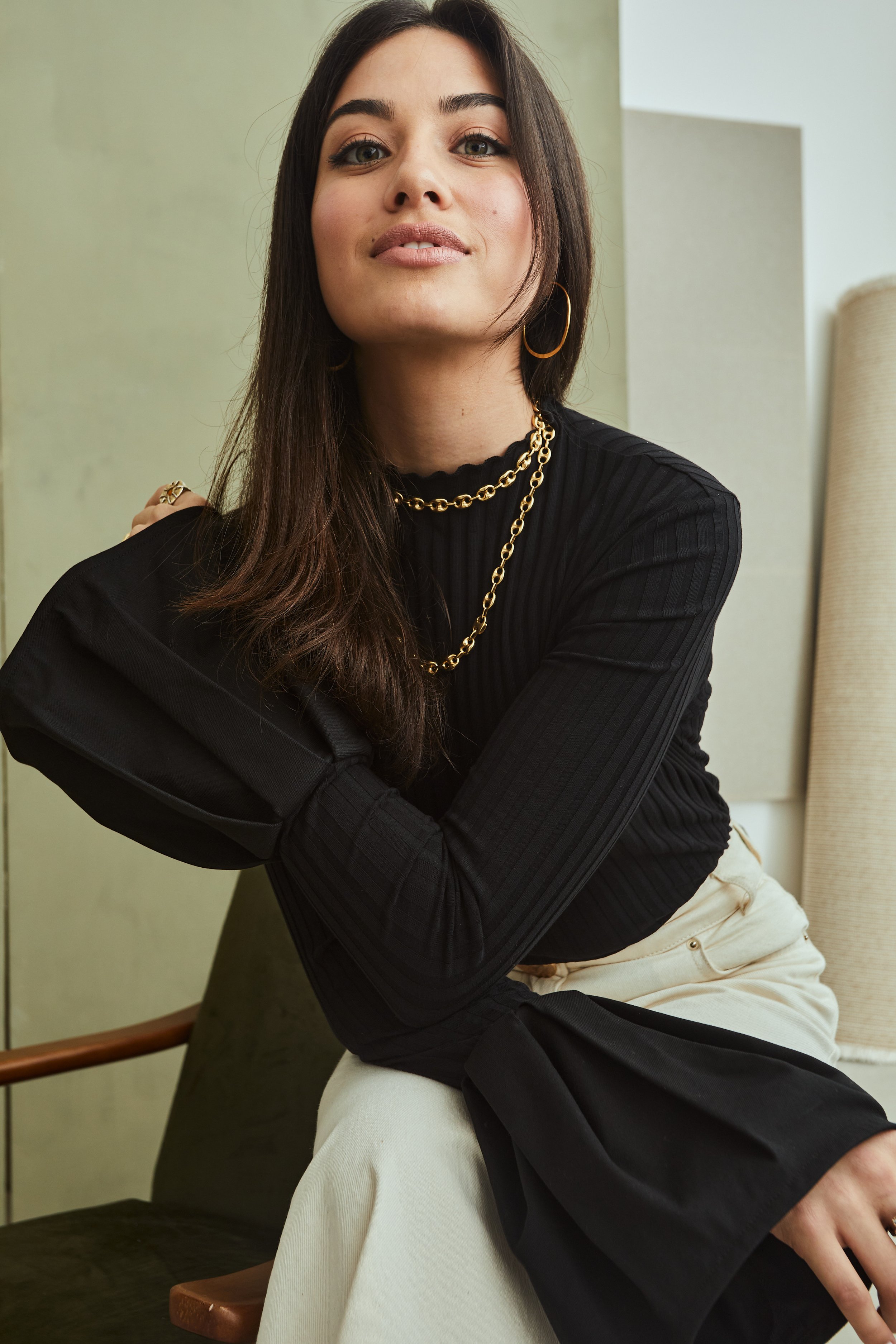 A young woman with long dark hair, wearing a black ribbed long sleeve top, beige pants, gold jewelry including earrings and layered chains, sitting on a chair with a neutral background.