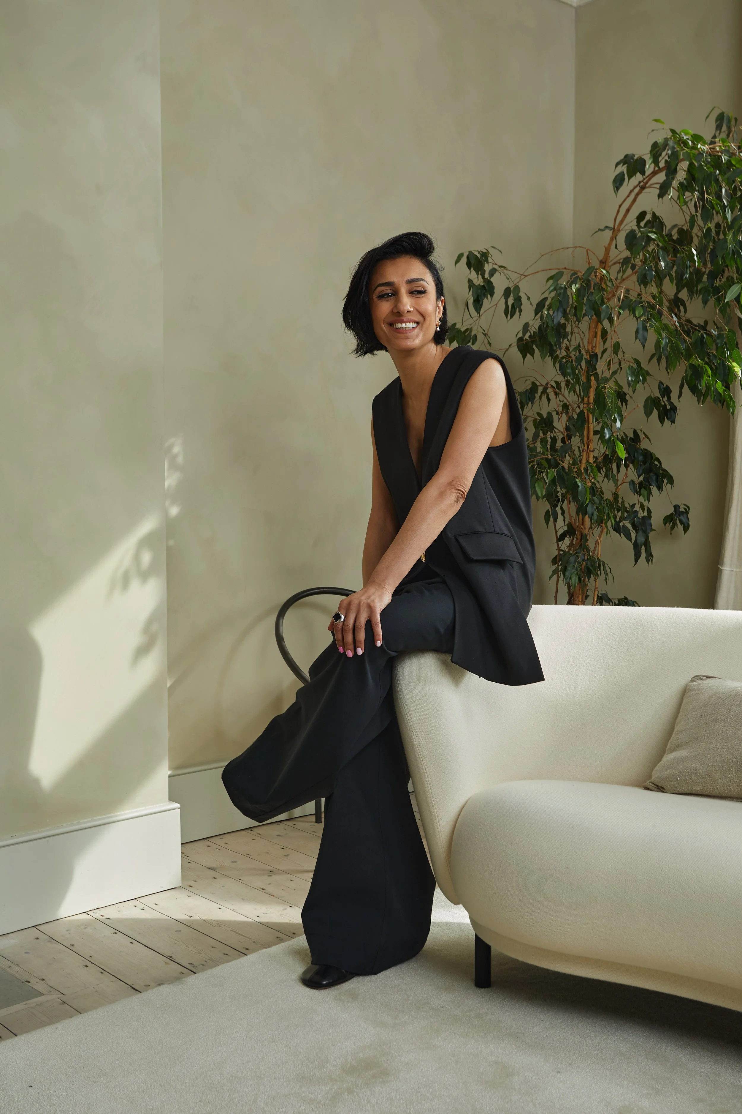Woman with short black hair smiling, sitting on a cream-colored sofa in a room with green walls and wooden floor, wearing a black sleeveless top and black pants, next to a large green plant.