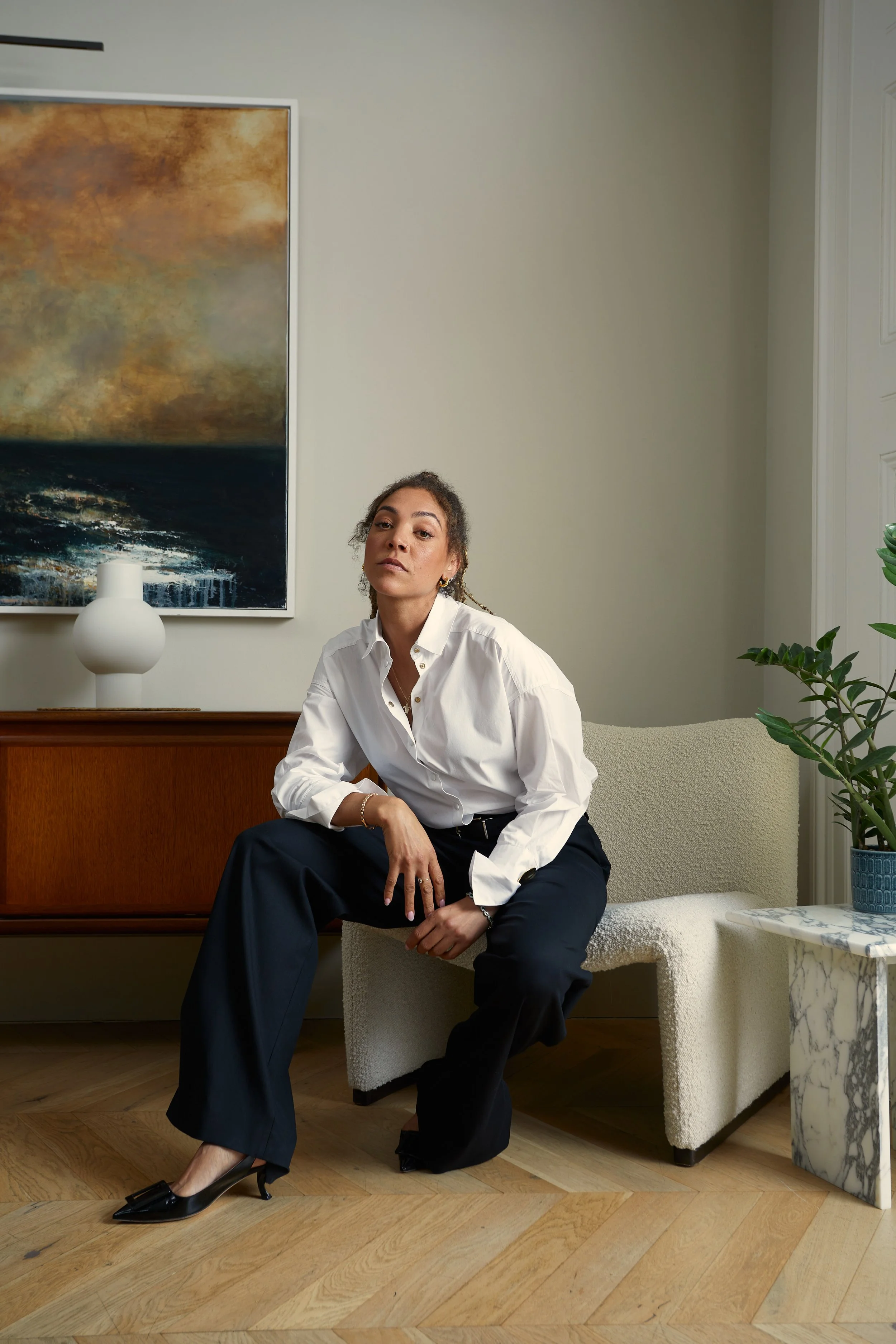 A woman sitting on a white textured chair in a modern living room, wearing a white shirt, black pants, and black heels, with a large painting, a white vase, and a potted plant in the background.