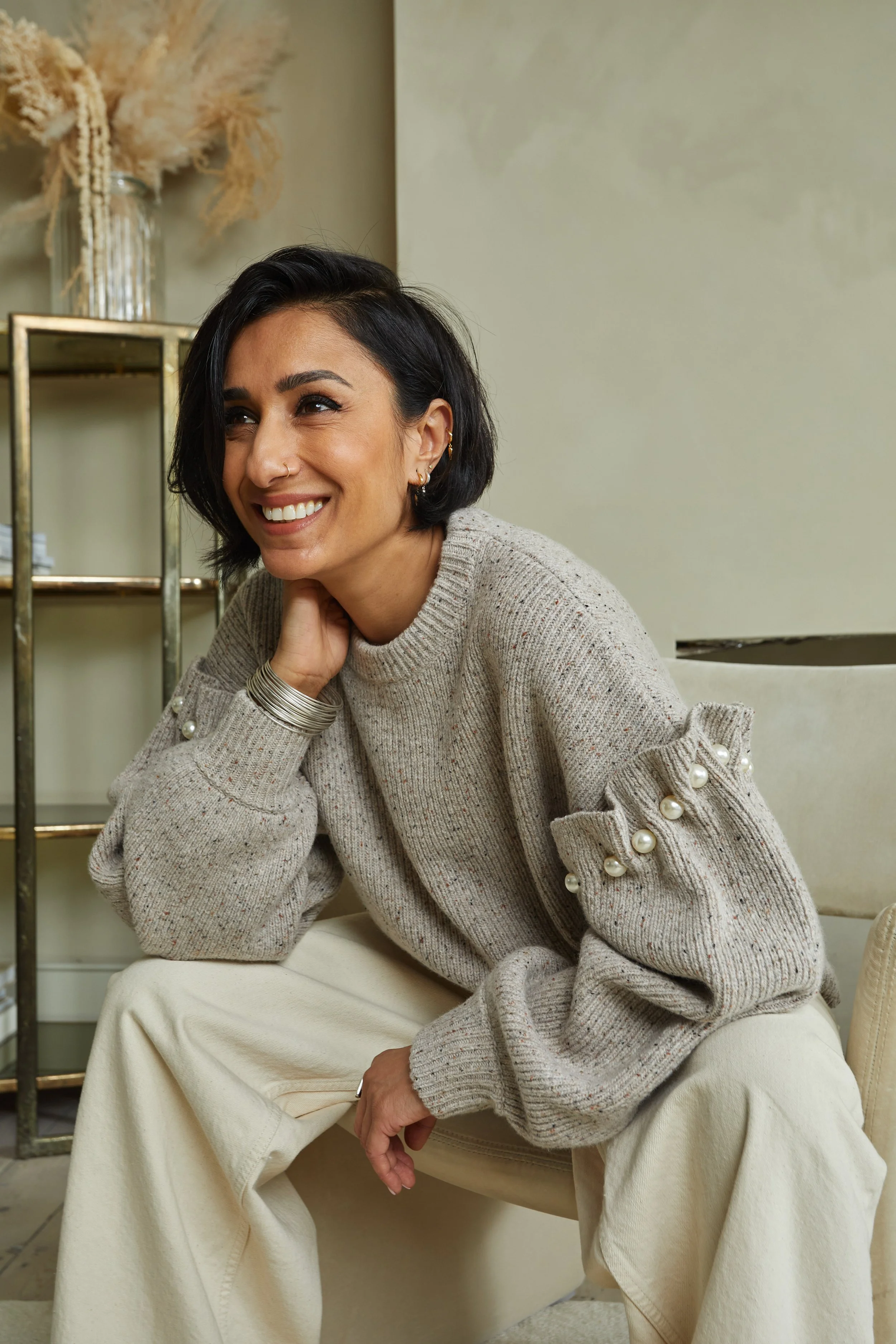A woman with short black hair, wearing a beige sweater with pearl embellishments on the sleeves, is sitting on a cream-colored chair in a room with neutral decor, smiling and looking to her right.