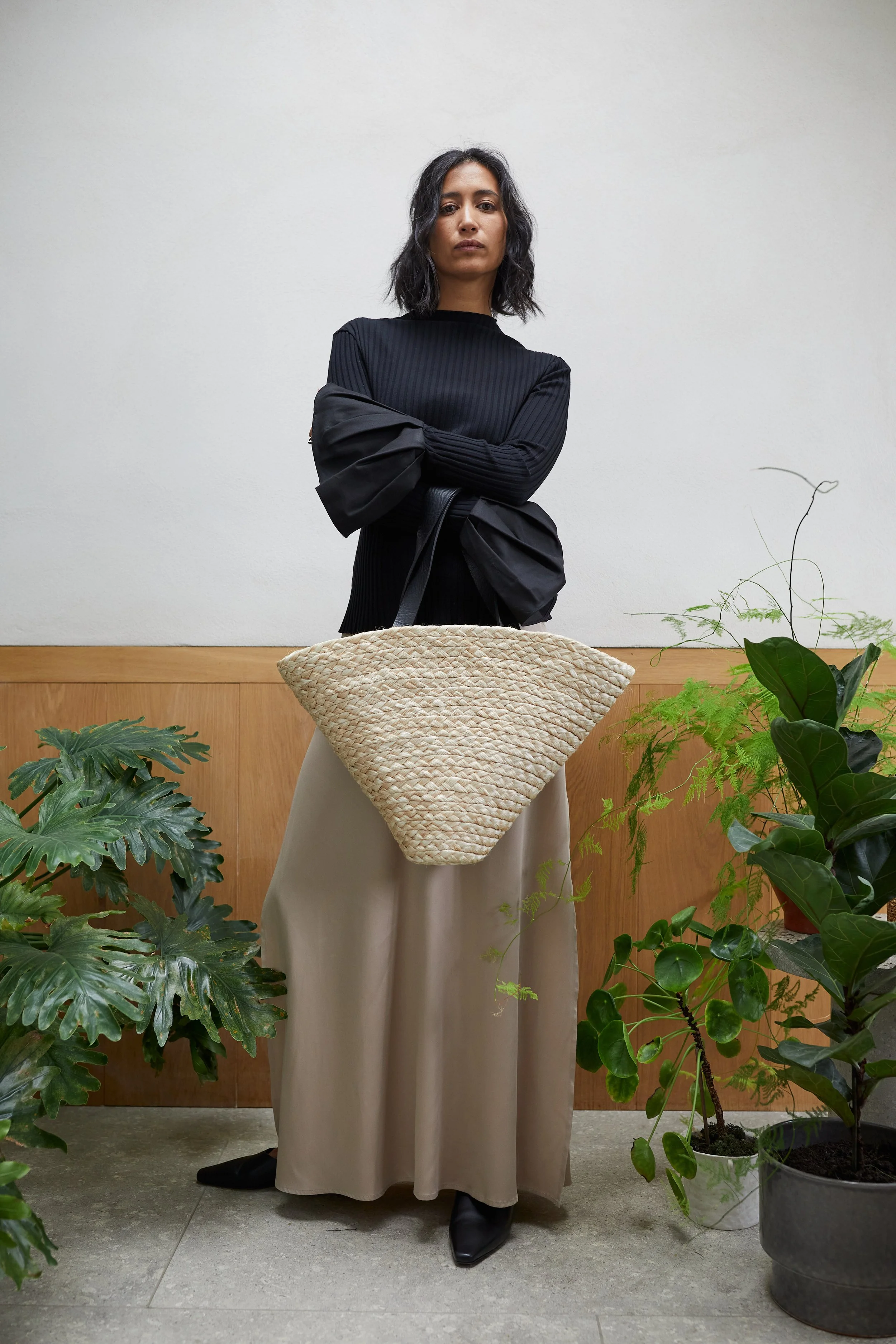 A woman in a black sweater and beige skirt standing behind a large woven tote bag, surrounded by green plants, against a plain white wall.