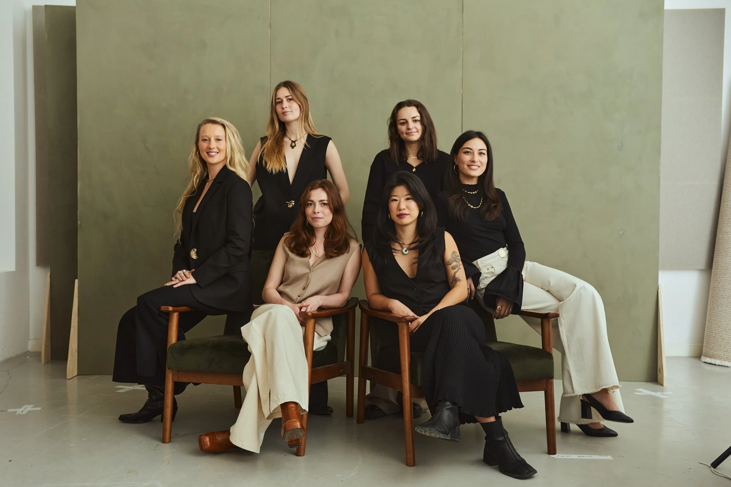 Group portrait of seven women posing together in front of a green backdrop, sitting and standing in fashionable black and beige outfits.