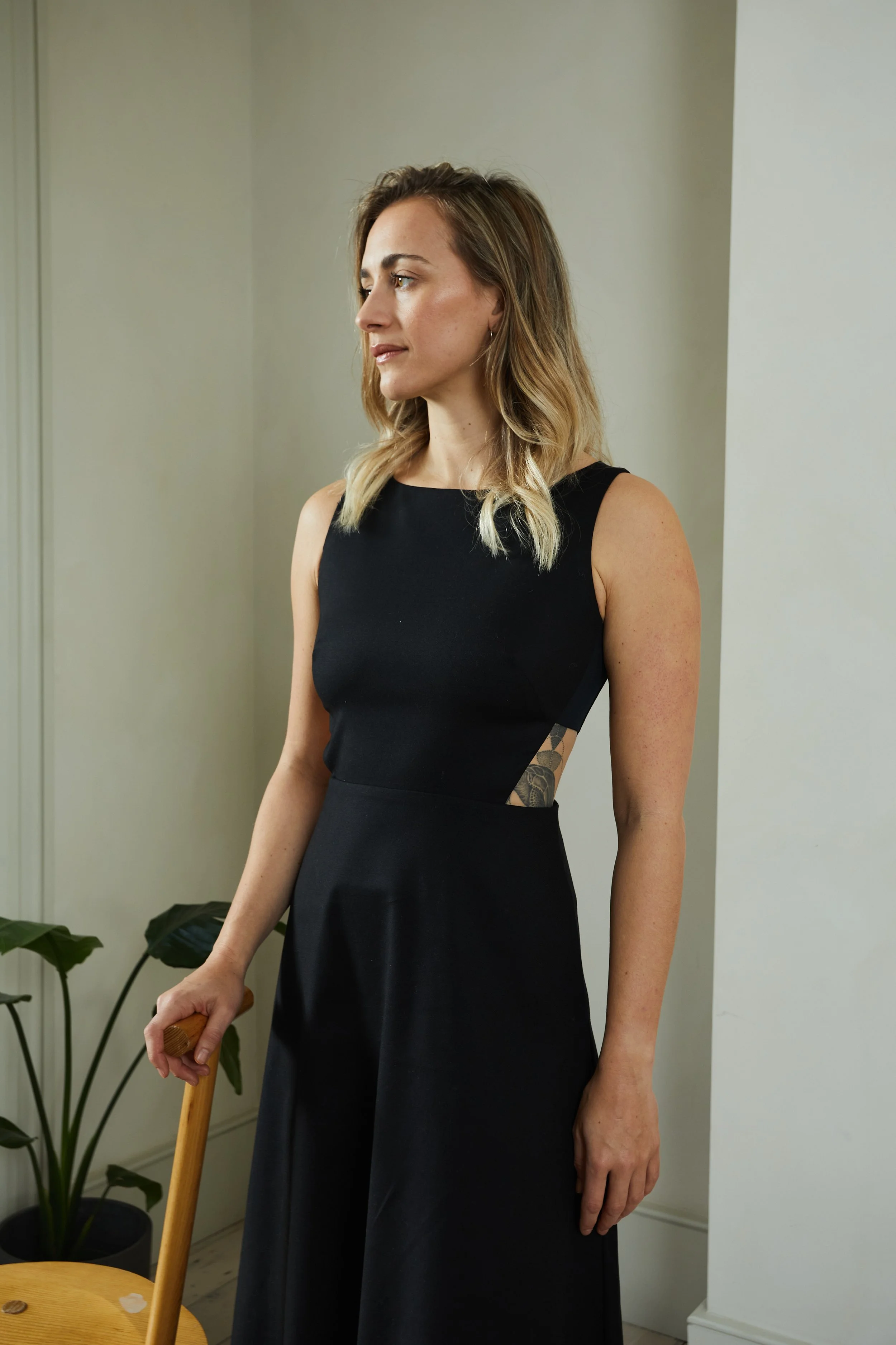 A woman with shoulder-length wavy blonde hair wearing a sleeveless black dress, standing indoors near a white wall, holding a wooden chair, with a houseplant nearby.