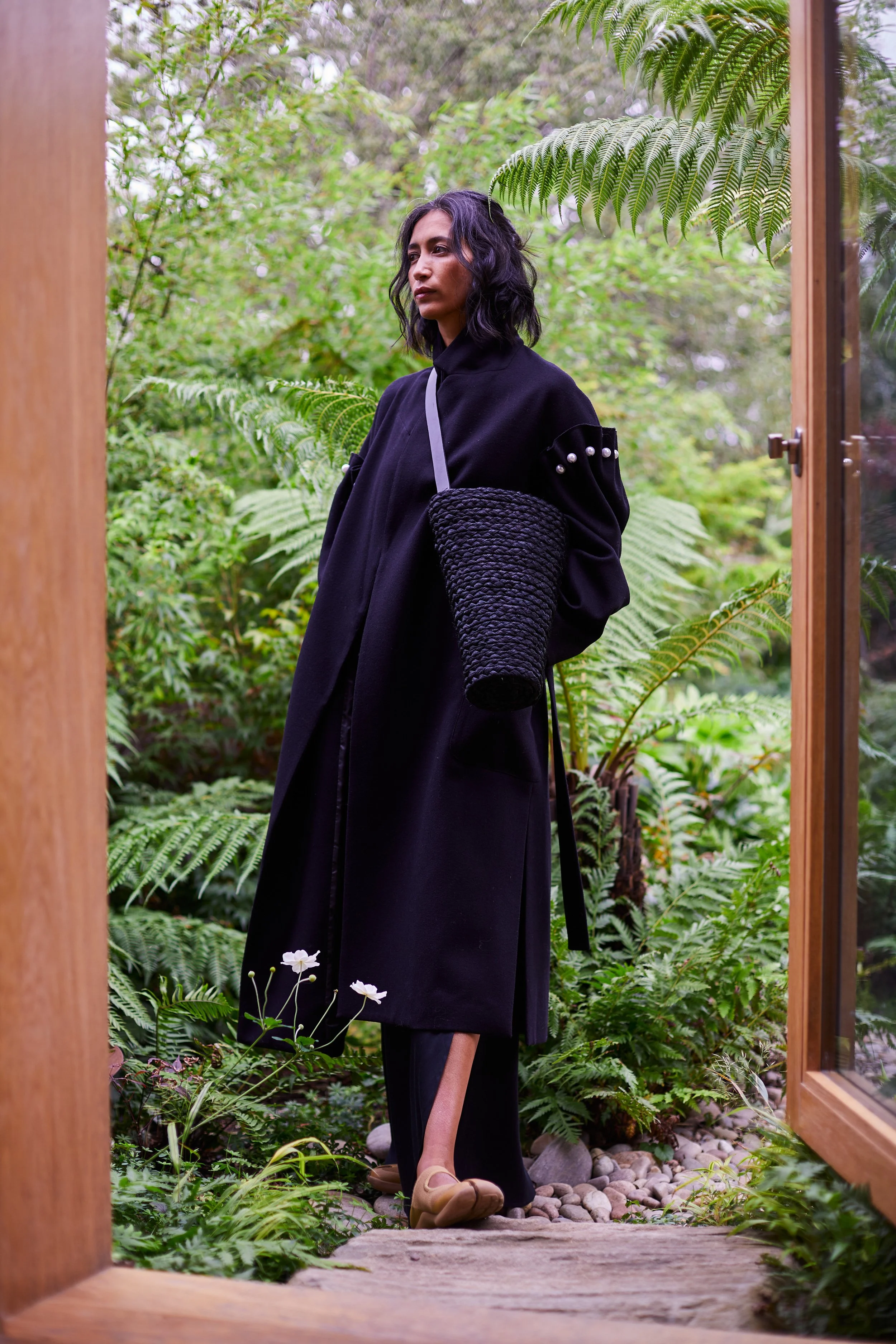 A woman with dark, wavy hair wearing a long black coat and beige shoes stands among lush green plants and ferns, viewed through an open wooden door.