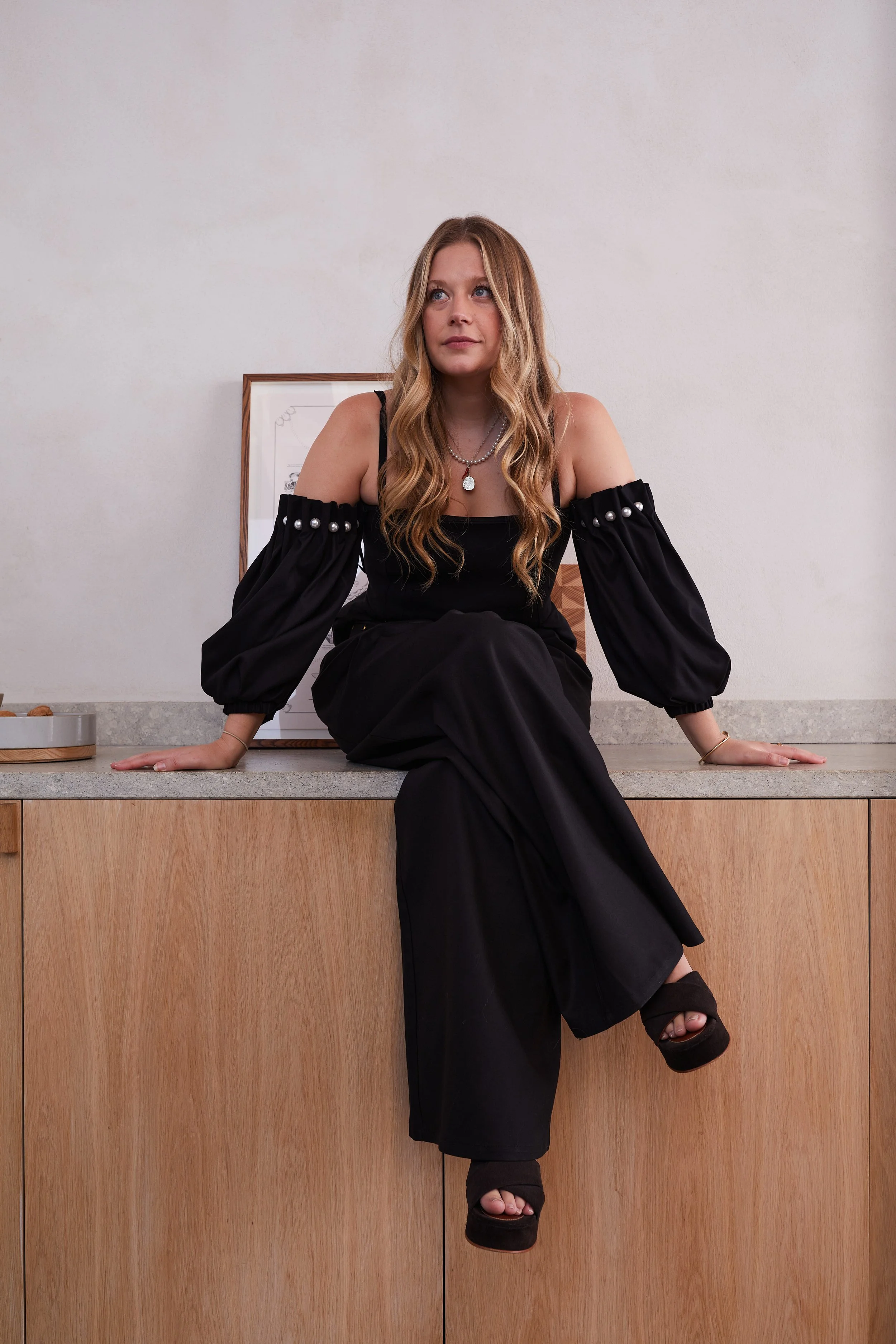 A woman with long wavy blonde hair sitting on a kitchen counter. She is wearing a black off-shoulder dress with puffy sleeves and pearl embellishments and black platform sandals. Behind her is a framed picture and a grey wall.