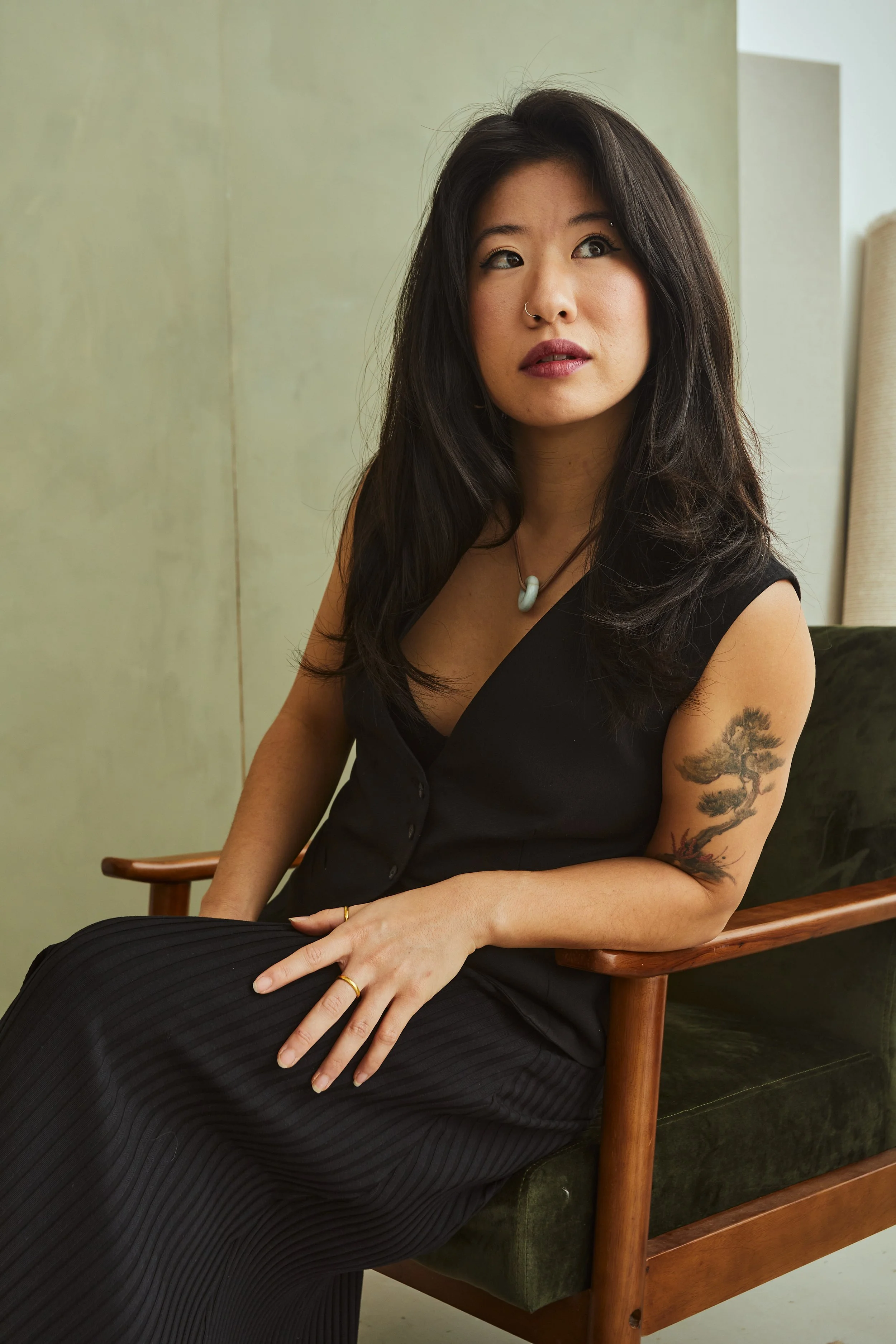 A woman with long dark hair wearing a black sleeveless top and black pants, sitting on a green couch with wooden armrests, looking to the side with a contemplative expression, in a room with green walls.
