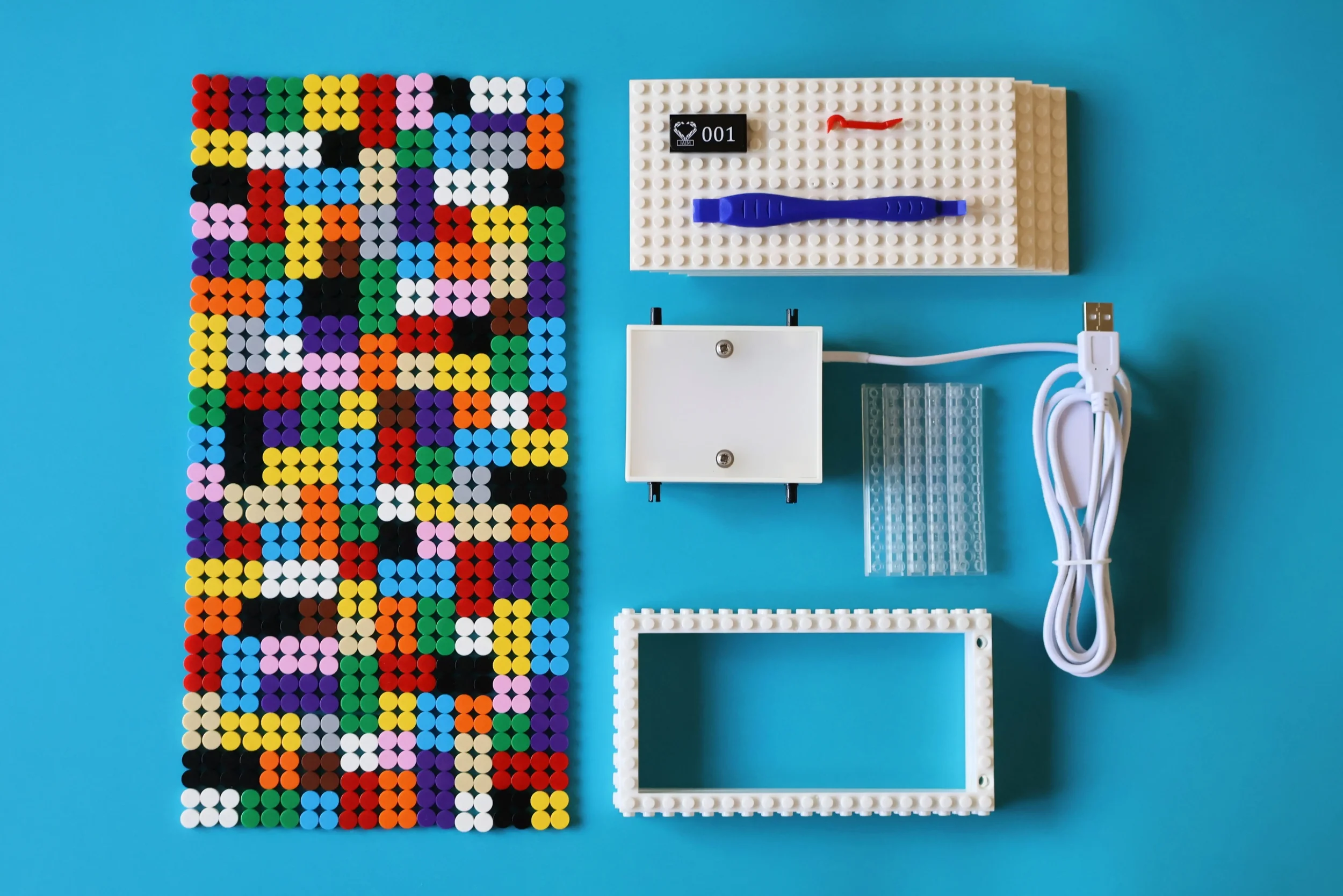 Colorful mosaic of small round beads, various shapes and colors, next to a plastic LEGO-like white display frame, a white plastic box with screws, a white LED light strip, a white power cord with a USB connector, a LEGO-like white panel with studs, and a white LEGO plate with studs on a bright blue surface.