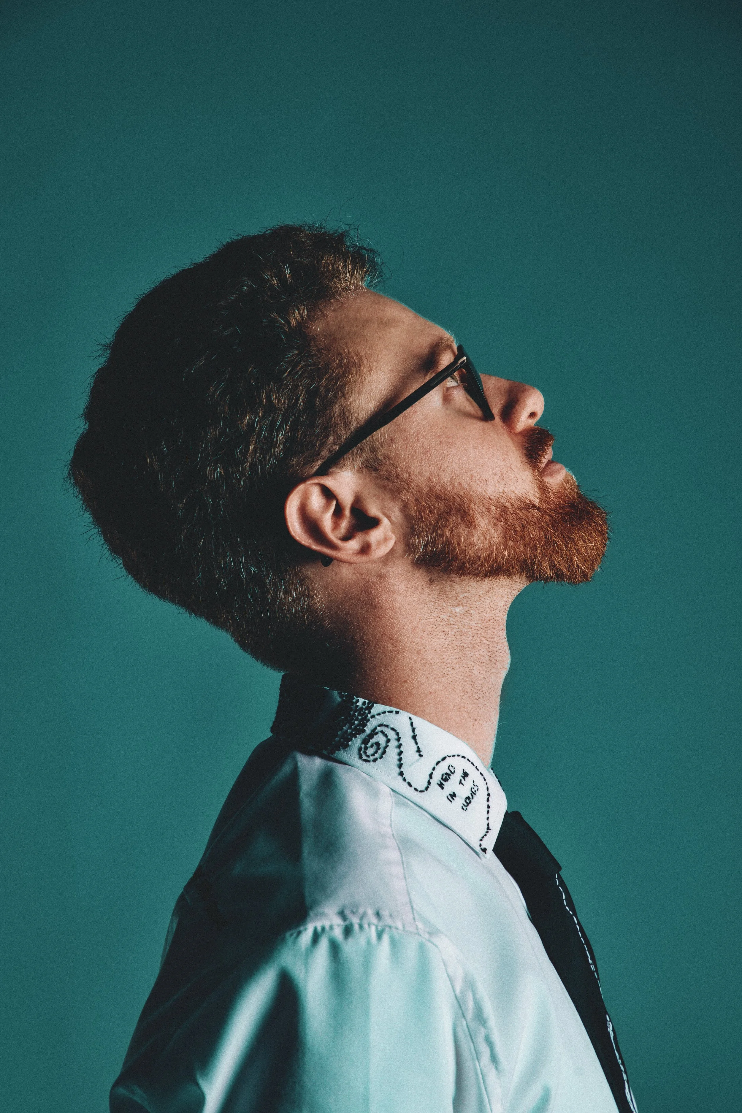 Side profile of a bearded man with glasses and styled hair wearing a white jacket with decorative black text and patterns, against a teal background.