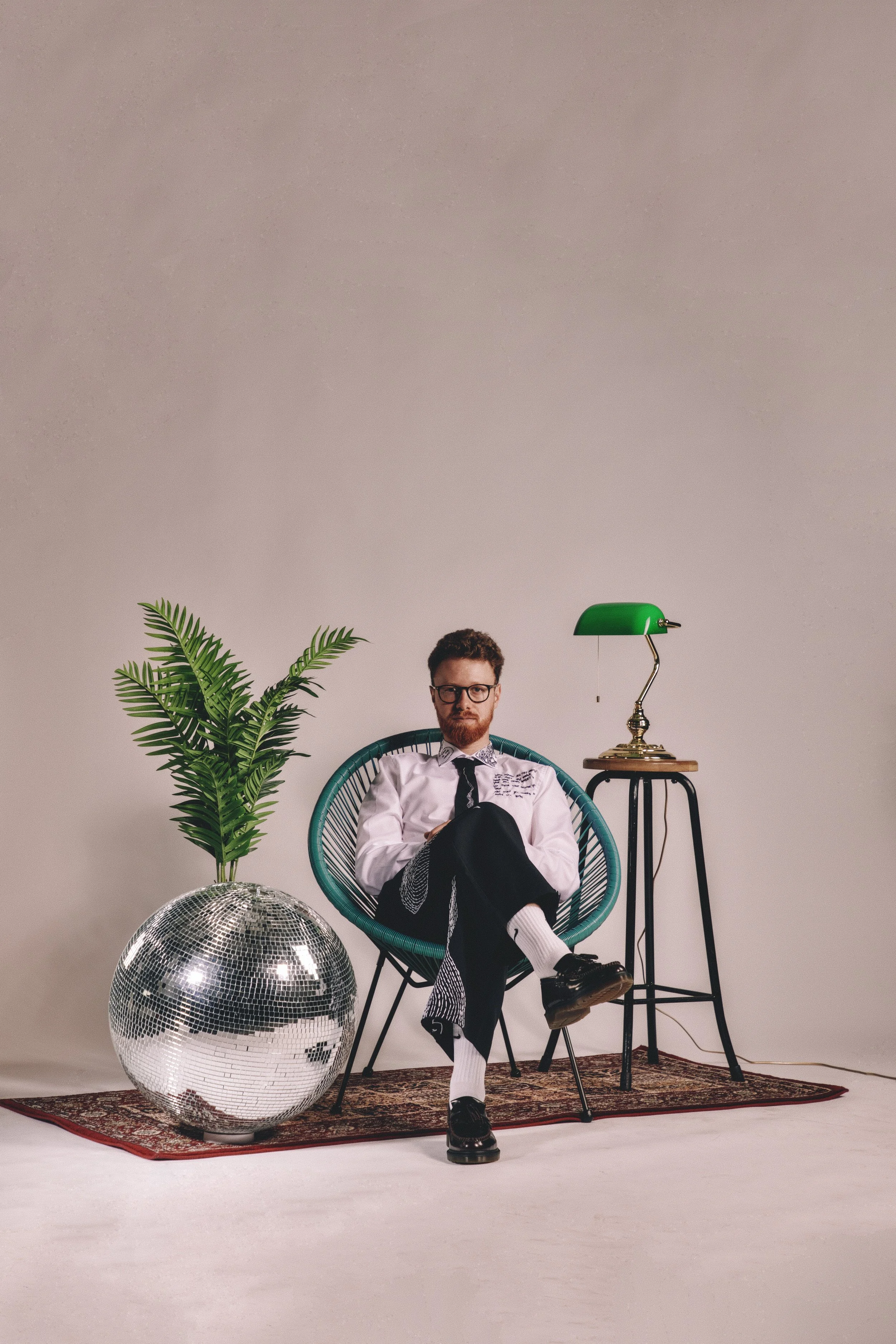 A man with curly hair and glasses sitting in a teal rounded chair, dressed in a white shirt, black tie, and black and white checkered pants. The room has a large disco ball on a red-patterned rug, a green desk lamp, and a large indoor plant, all agai
