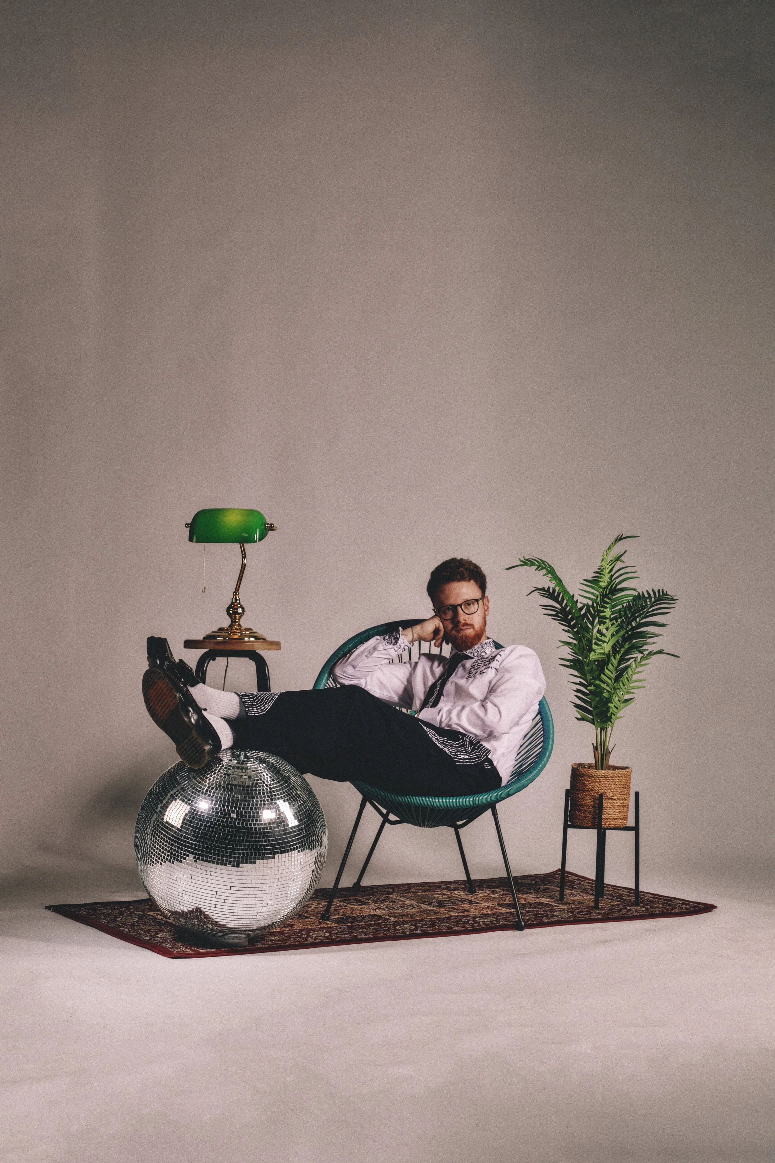 A man with glasses, a beard, and a mustache, sitting in a green chair, wearing formal attire with a white shirt, black tie, and black pants with white patterns, resting his head on his hand and looking at the camera. There is a disco ball on the floo