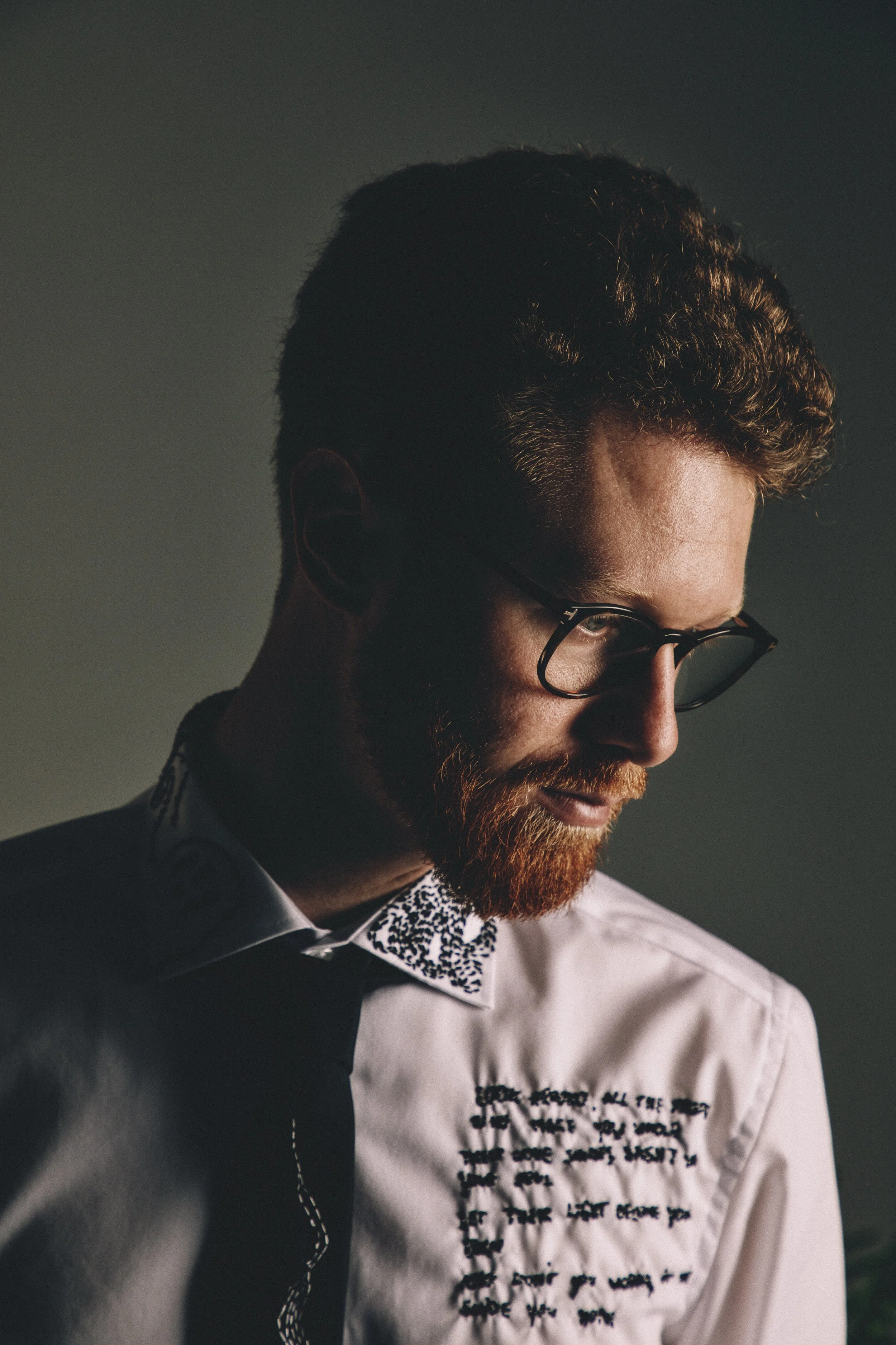 A man with a beard wearing glasses and a white shirt with black text and patterns, looking down, with side lighting against a dark background.