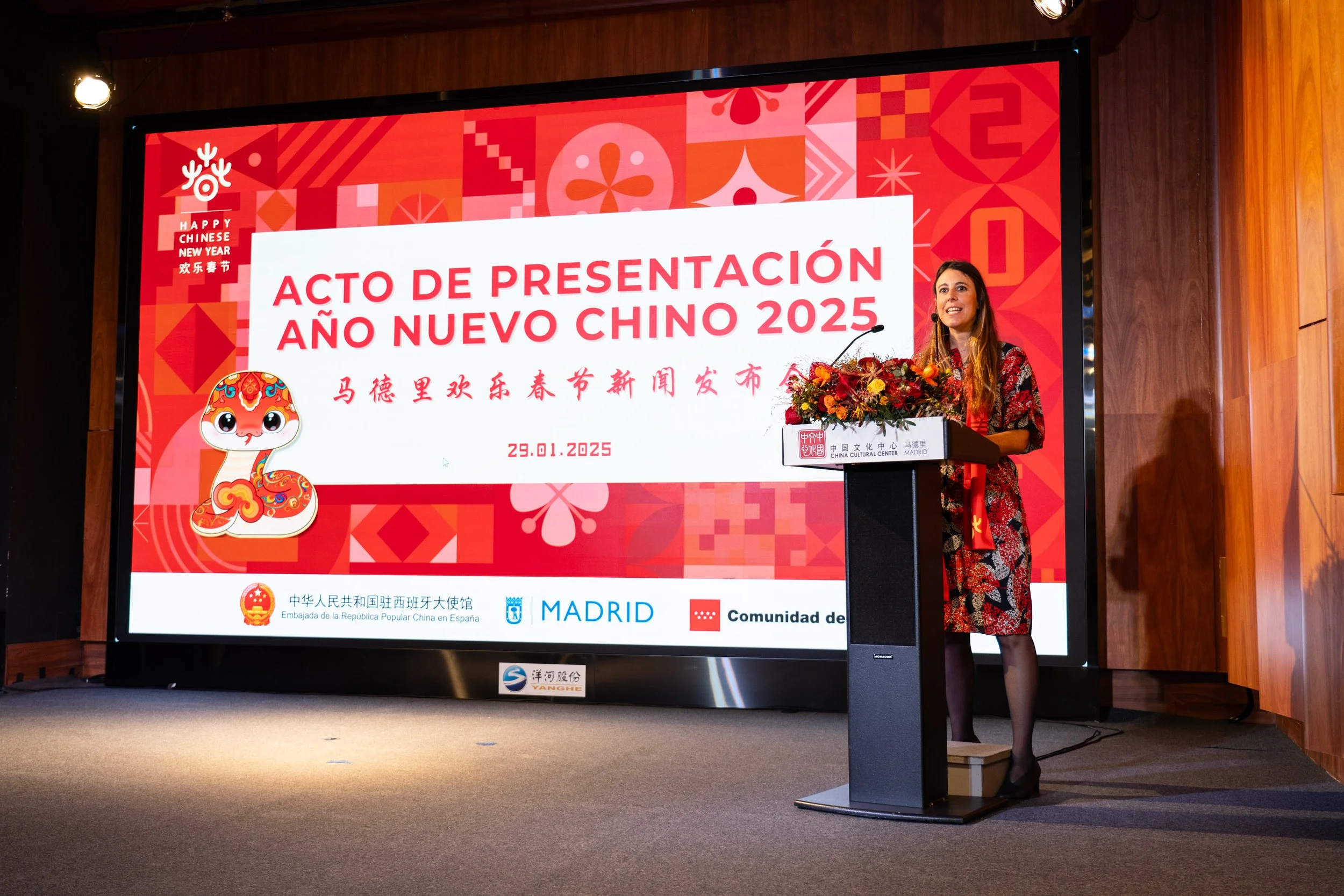Mujer hablando en un evento en un escenario con un gran cartel que dice 'Acto de presentación Año Nuevo Chino 2025'.