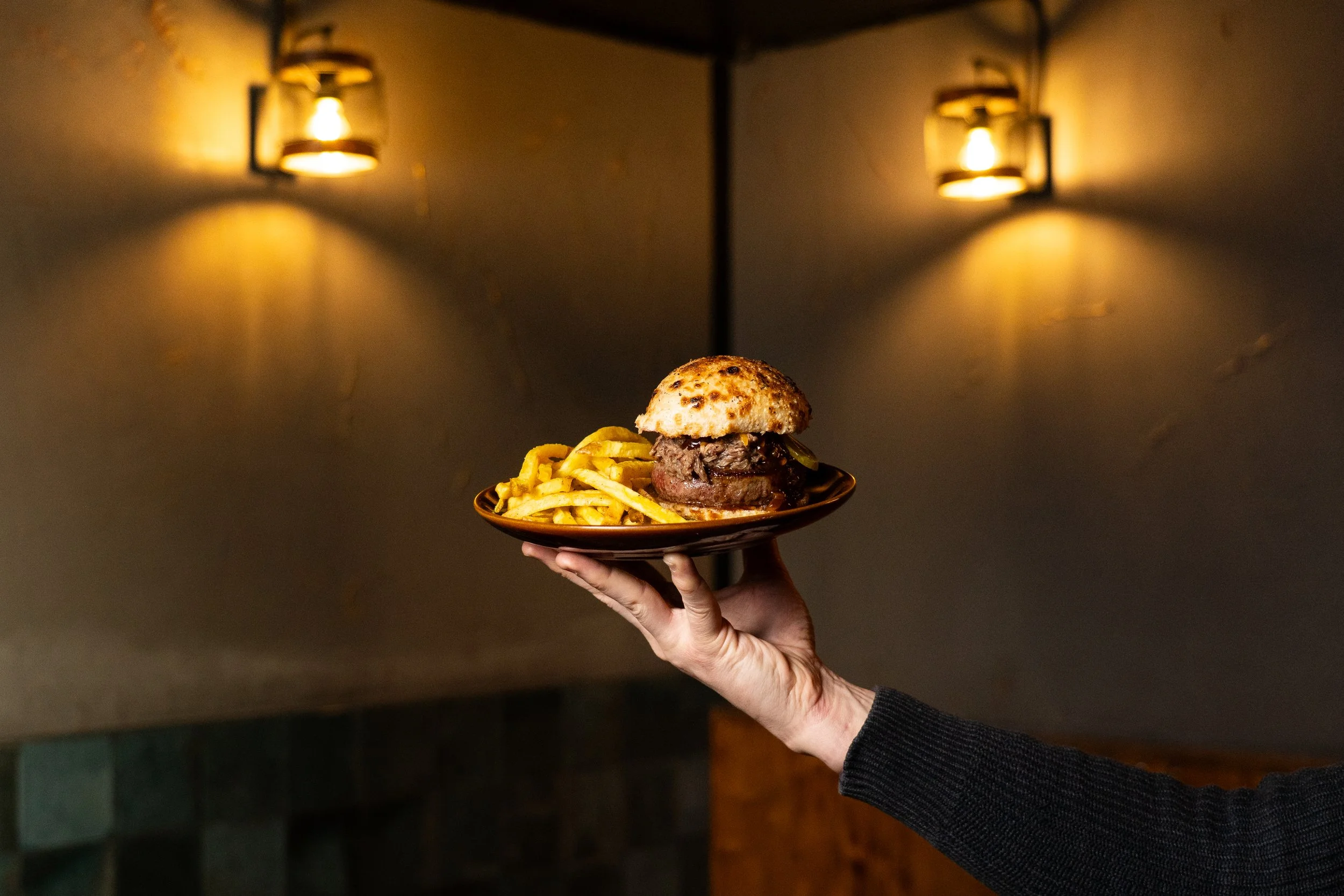 Una mano sostiene un plato con una hamburguesa y papas fritas en un fondo oscuro con luces cálidas en la pared.