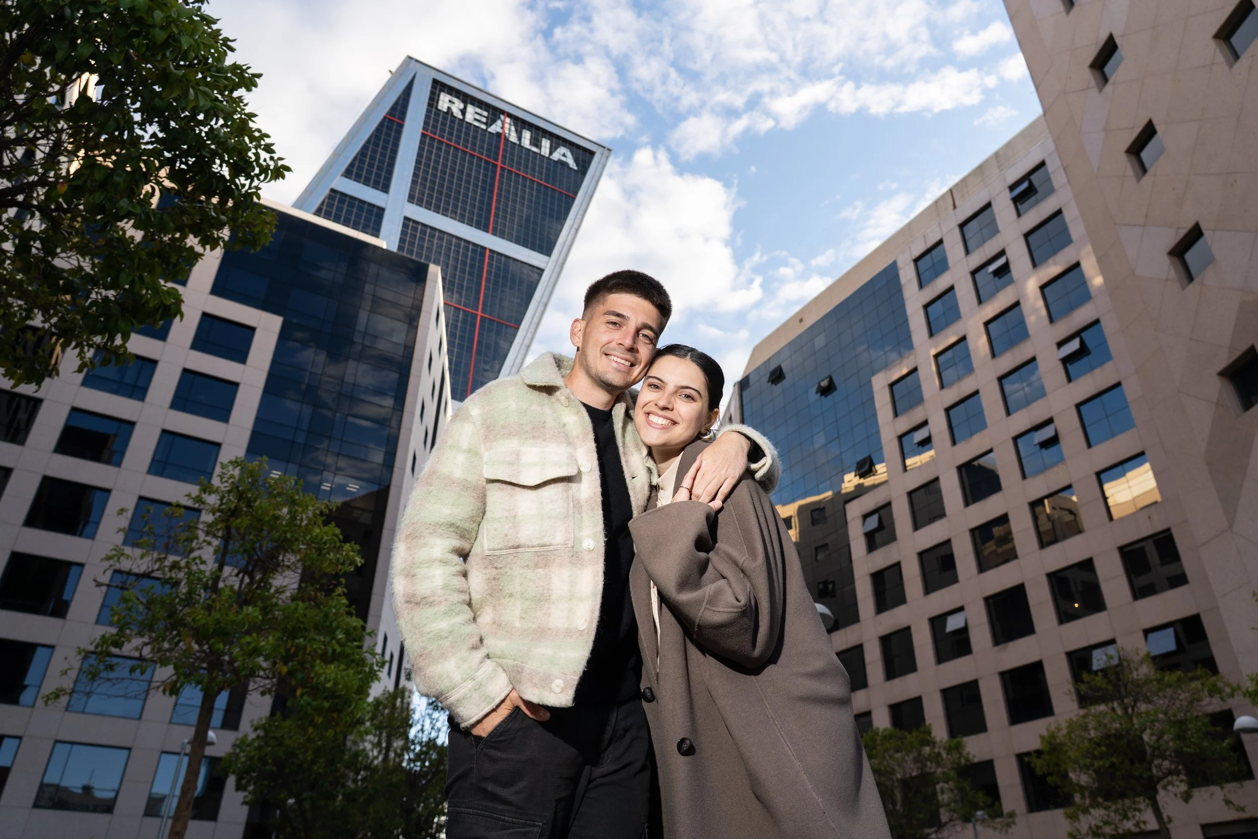 Una pareja sonriendo en la calle frente a edificios modernos con cielo nublado y un árbol a la izquierda.