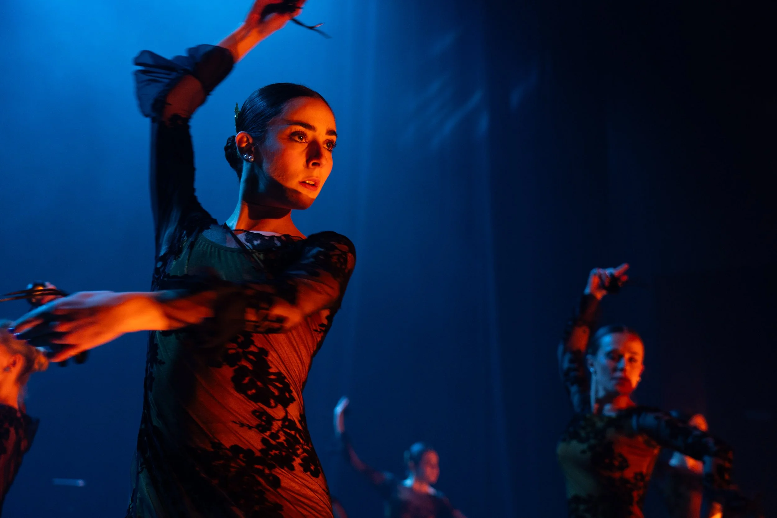Mujer bailando en un escenario con iluminación azul y naranja, vestida con ropa de danza con detalles de encaje negro, y con cabello peinado en un estilo elegante.