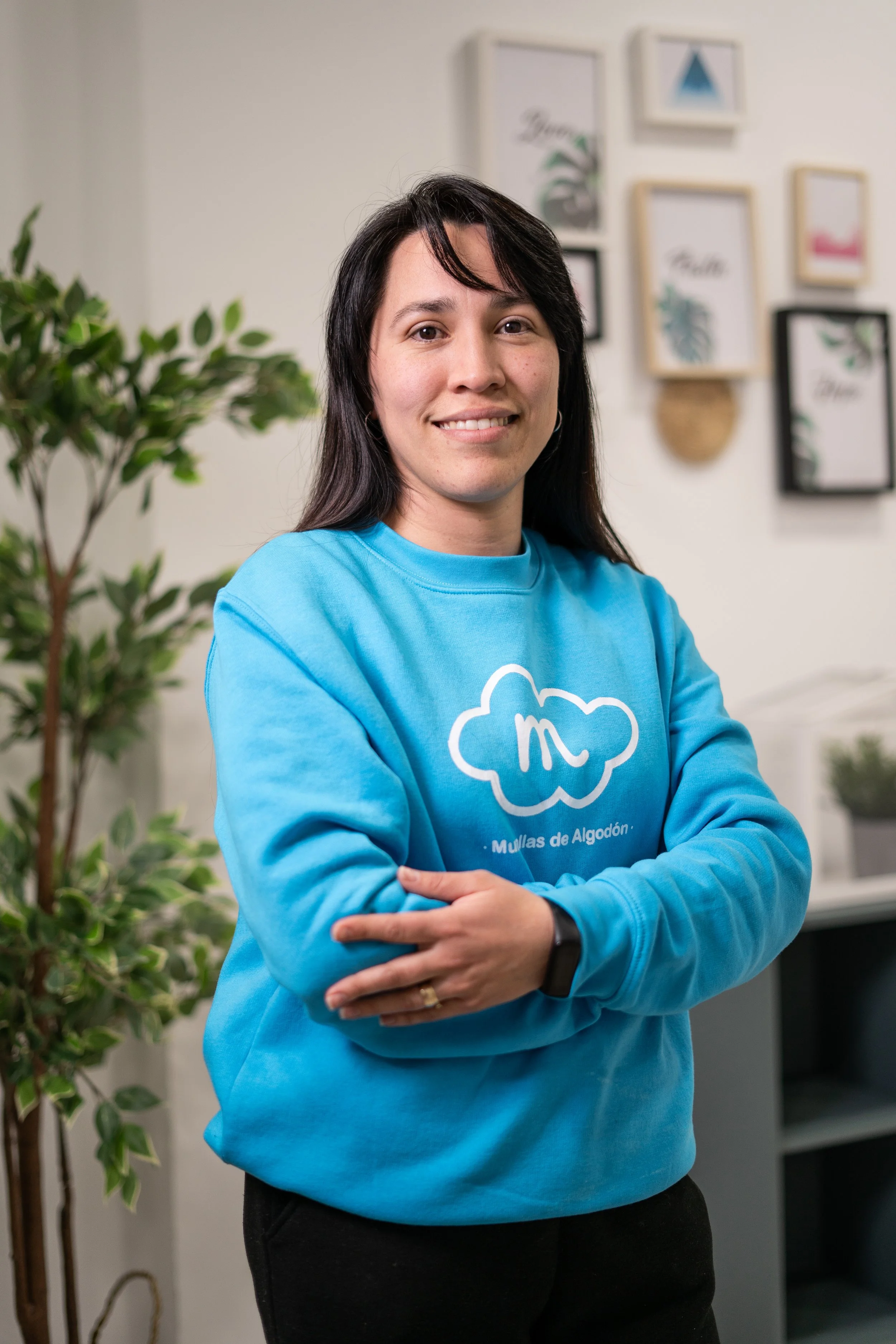Mujer de cabello oscuro, sonrisa y piel clara, lleva sudadera azul con logo de una nube y letras, de pie con brazos cruzados en interior decorado con cuadros y planta verde