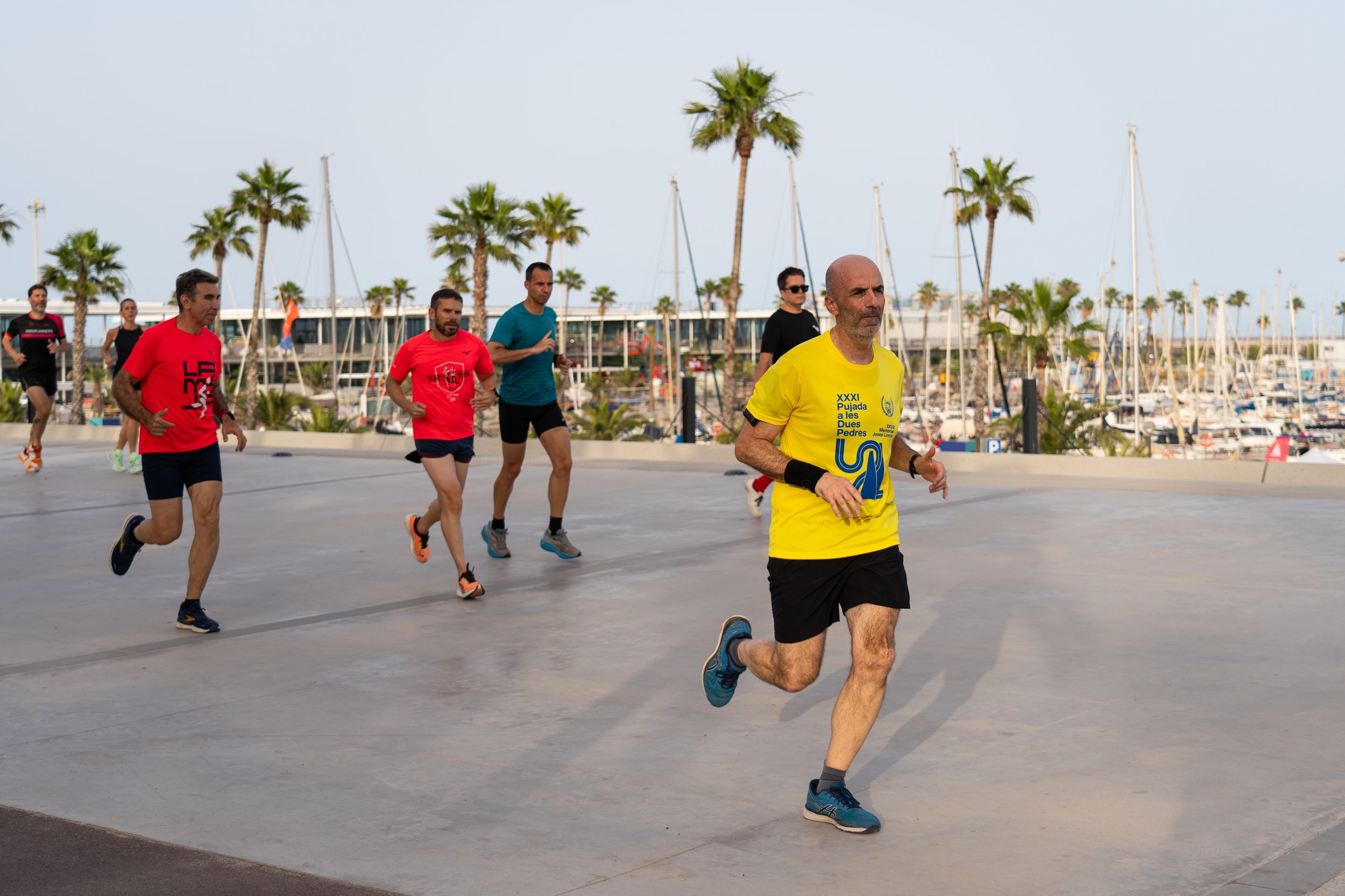 Hombres corriendo en un evento de maratón en un lugar con palmeras y mástiles de yates en el fondo.