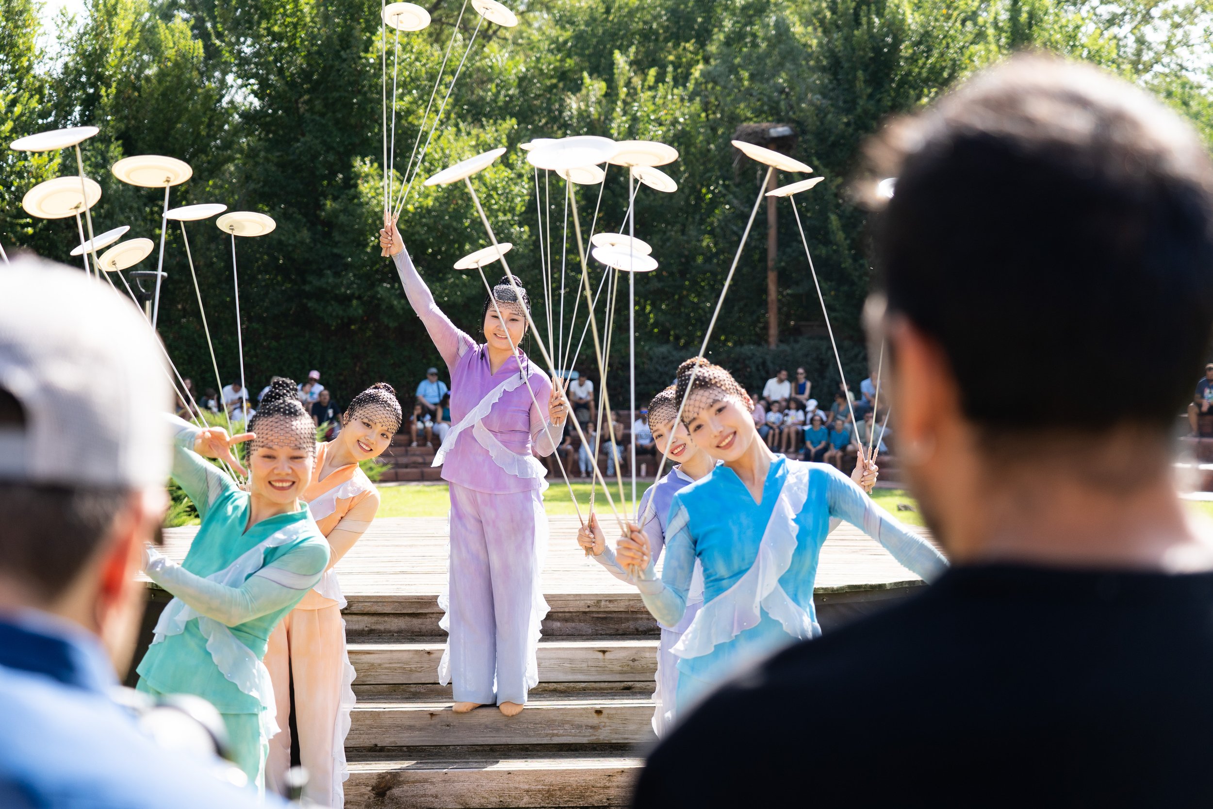 Grupo de bailarinas en un escenario al aire libre, vestidas con trajes de colores pastel, realizando una danza con sombrillas en un día soleado, con audiencia observando en el fondo.