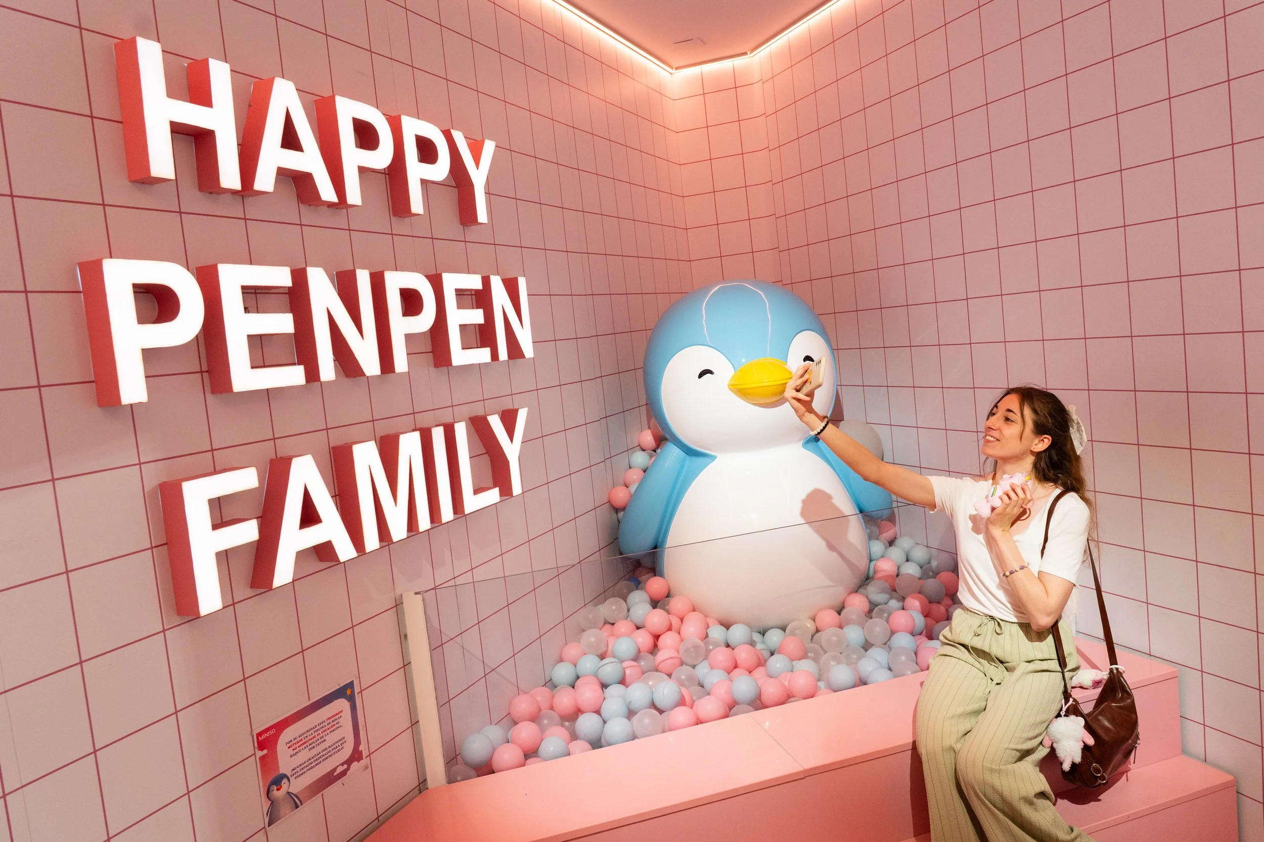 Una mujer con ropa a rayas sentada en el borde de una zona decorada con una figura de pingüino inflable, bolas en rosa, azul y blanco en el suelo, y letras en neón que dicen 'HAPPY PENGUIN FAMILY' en la pared. La mujer están sonriendo y tocando la figura del pingüino.