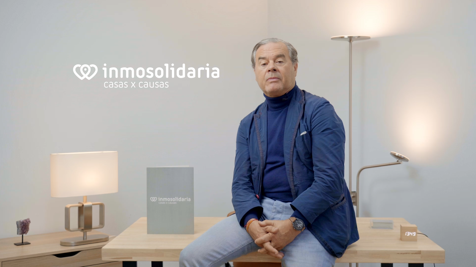 Hombre sentado en una mesa de madera con fondo gris, con logotipo y texto 'Inmosolidaria casas x causas', acompañado por una lámpara y otros objetos decorativos.