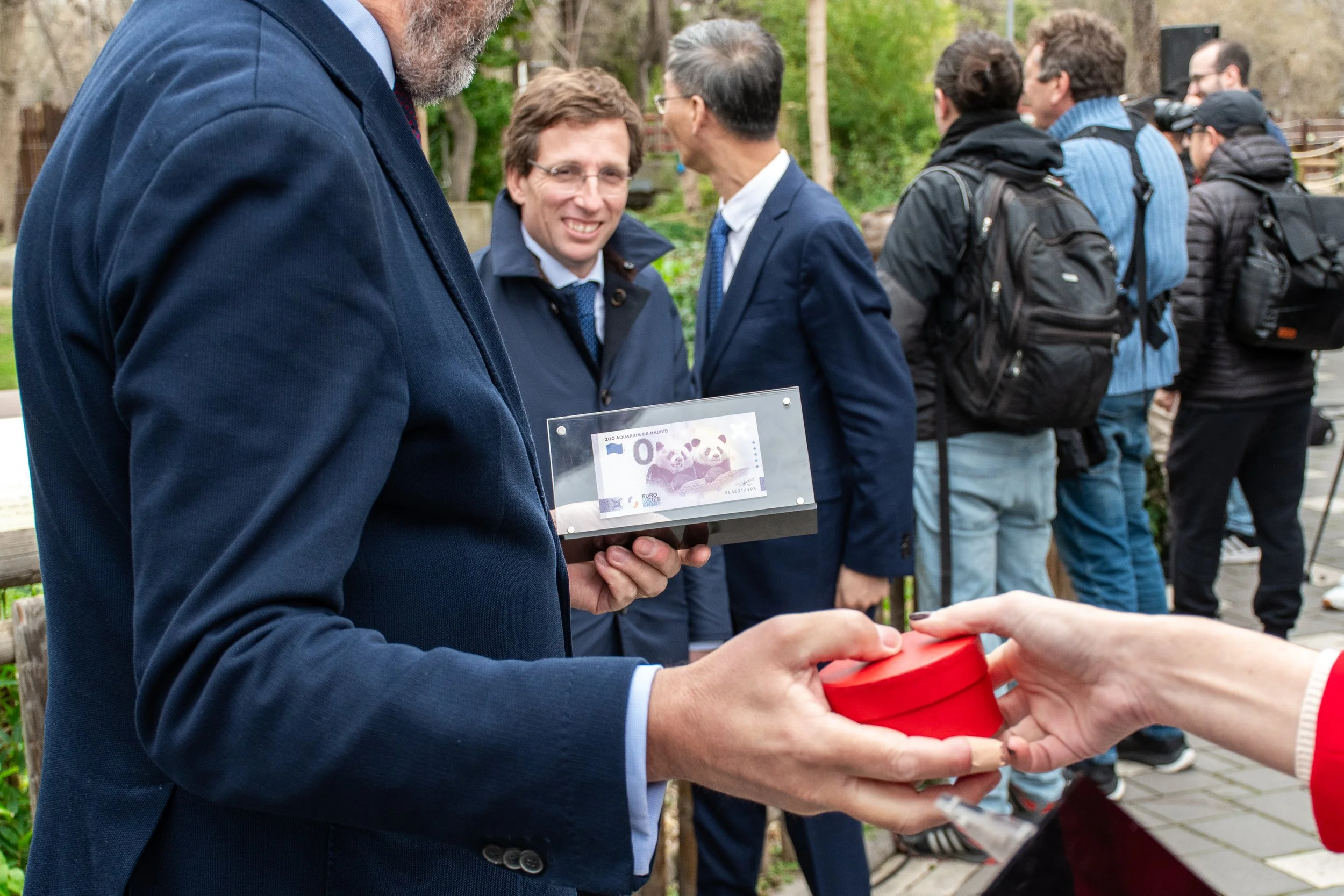 Un hombre vestido con traje y corbata recibe una caja de regalo de color rojo en un evento al aire libre con varias personas, algunas con mochilas, en un entorno natural.