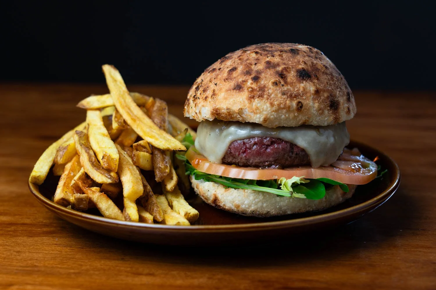 Hamburguesa con queso, tomate, lechuga, carne y pan, acompañada de papas fritas, sobre plato de cerámica en mesa de madera.