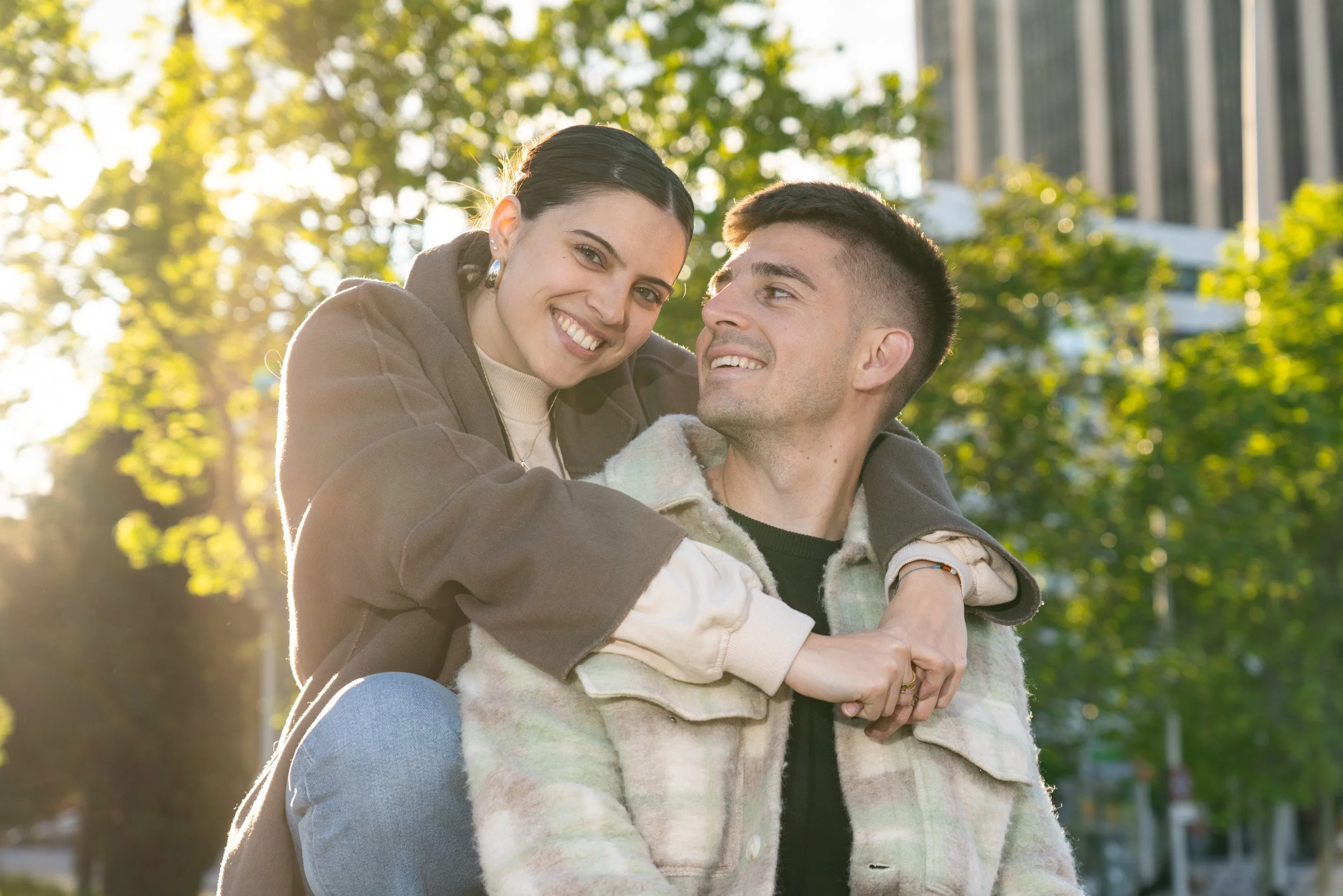 Una pareja joven sonriendo en un parque con árboles y edificios de fondo en un día soleado.