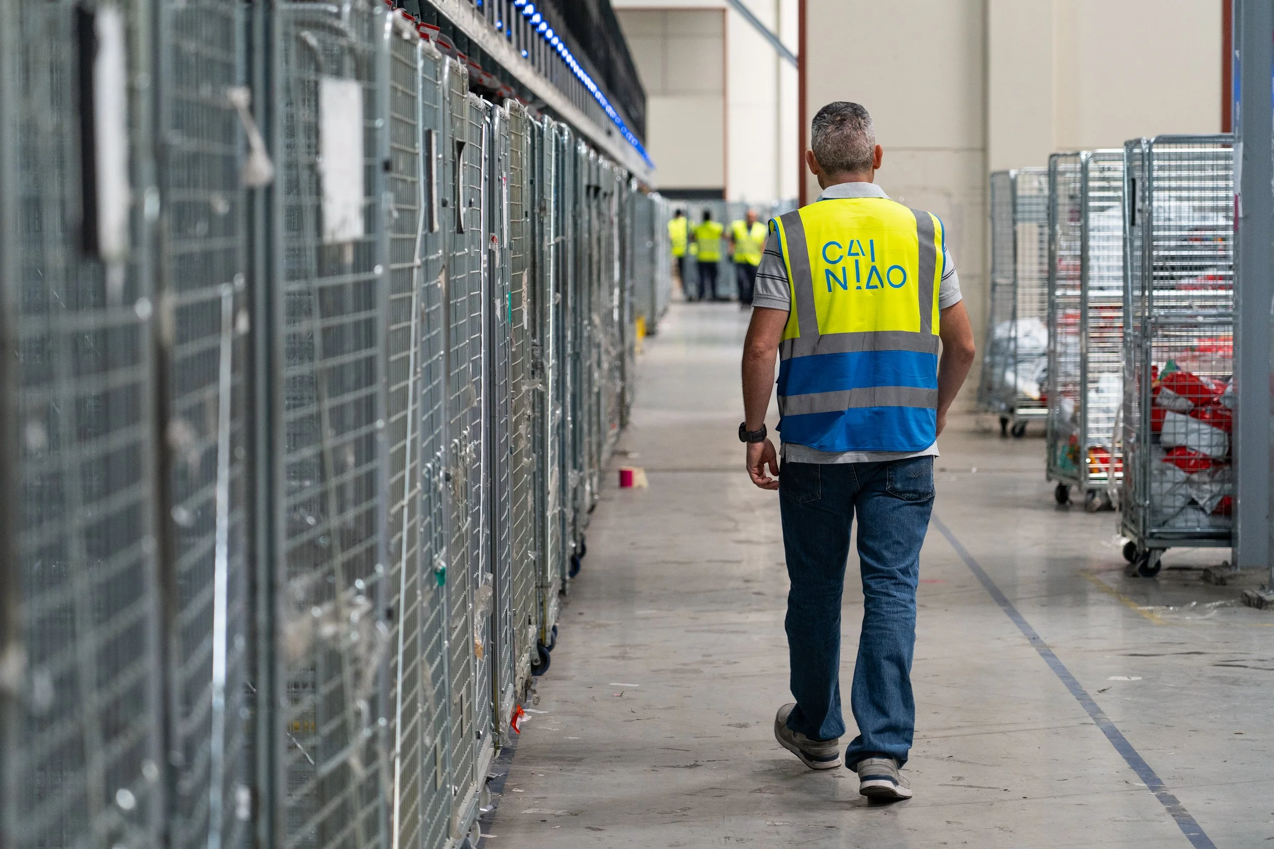 Una persona de espaldas, vistiendo un chaleco de seguridad amarillo con letras azules y pantalones vaqueros, caminando por un pasillo en un almacén con estanterías llenas de cestas metálicas, algunos empleados en el fondo.