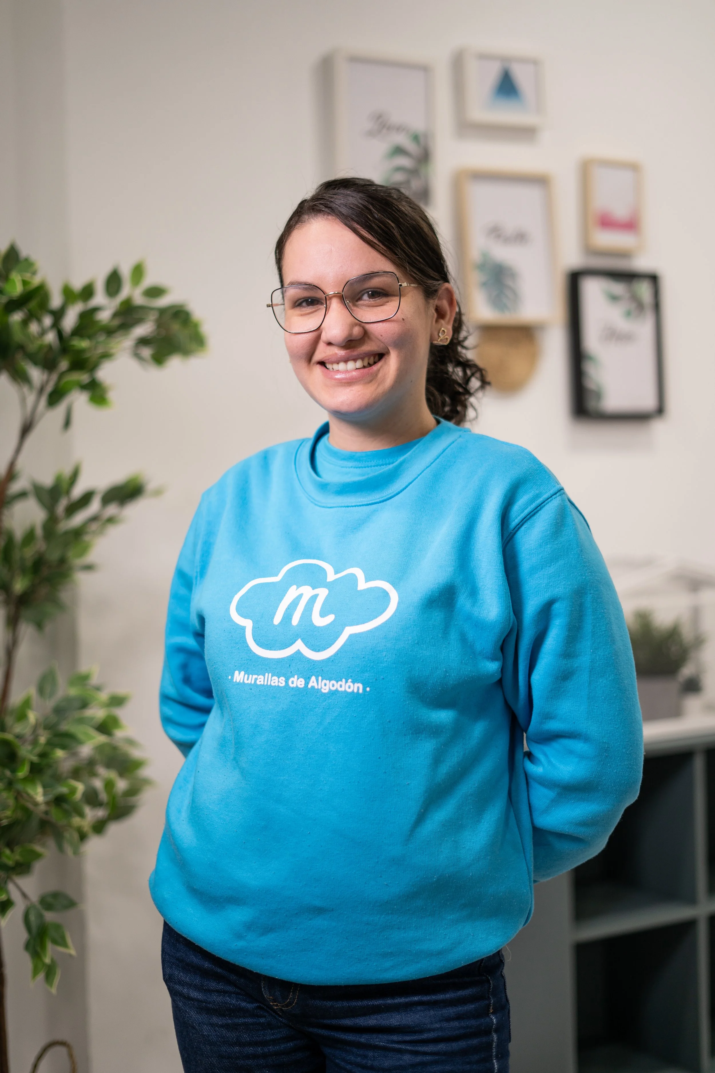 Mujer joven con gafas, camiseta azul con logotipo y texto en español, sonriendo en interior con cuadros decorativos en la pared y planta en primer plano.