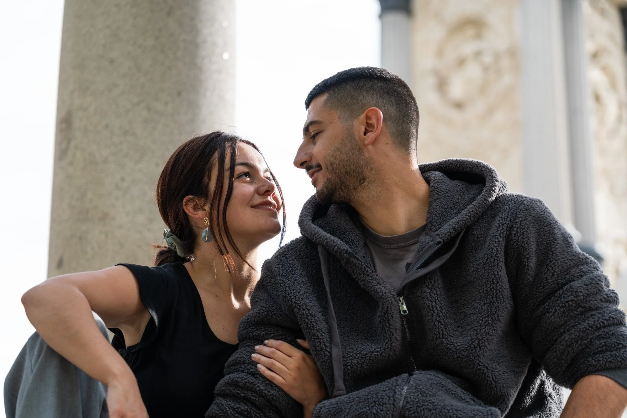 Una pareja joven con expresión cariñosa, ellos se están mirando en una escena romántica, con fondo de columnas y arquitectura clásica.
