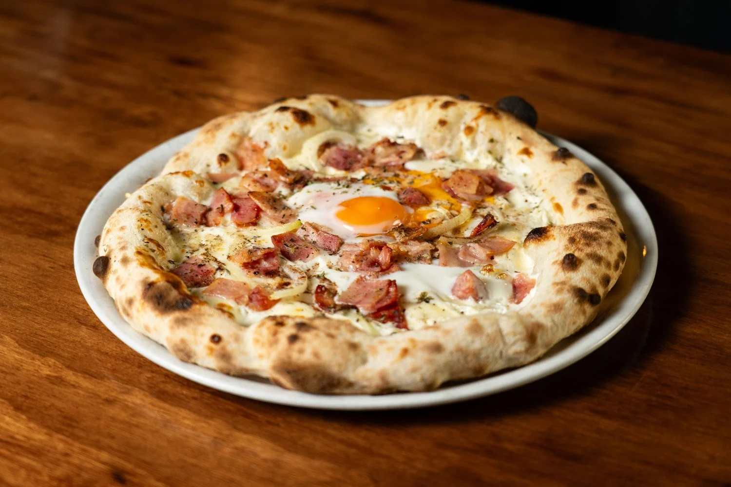 Pizza con huevo, jamón y tocino en plato blanco sobre mesa de madera.