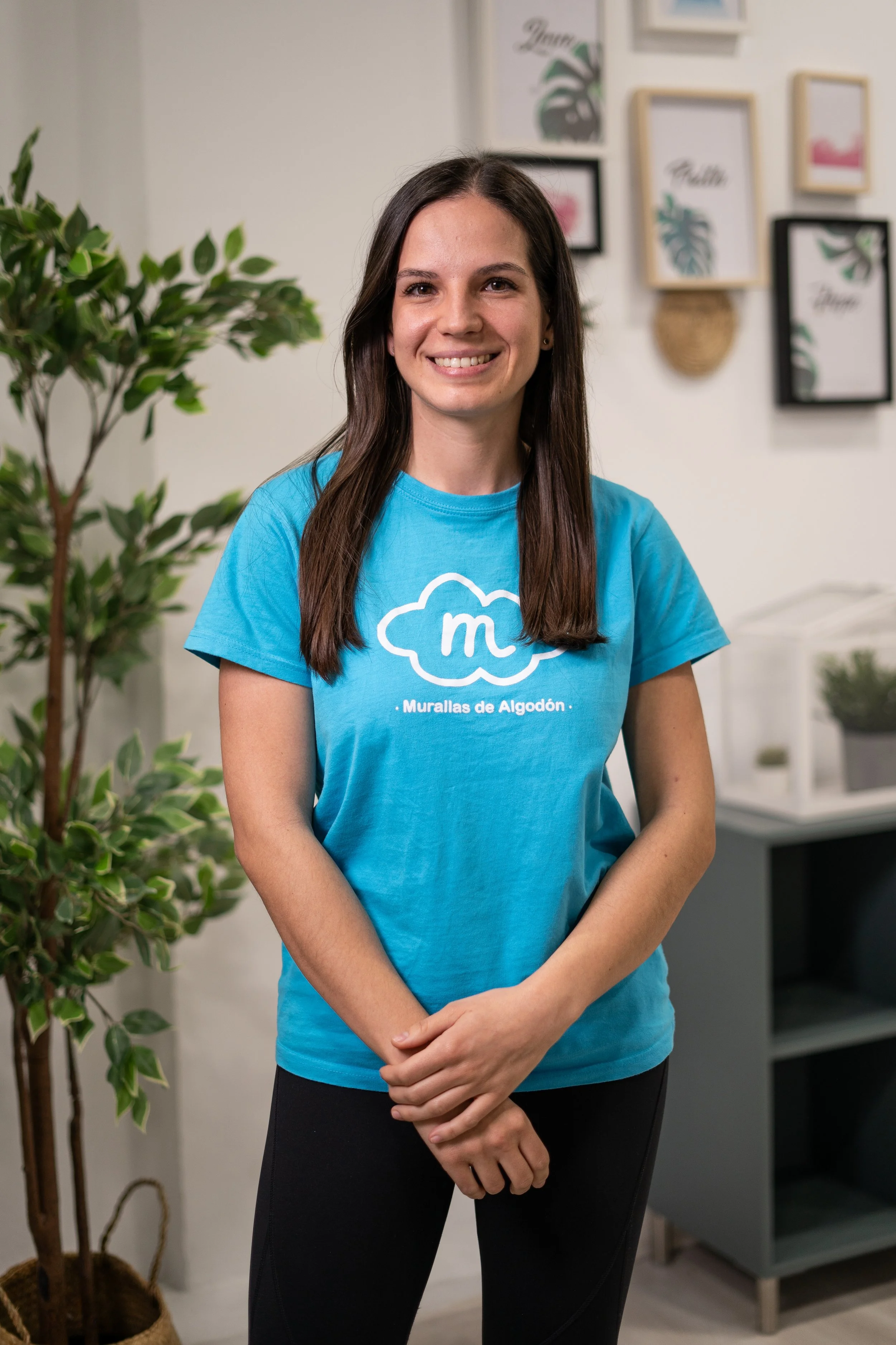 Mujer joven sonriendo, usando camiseta azul con logo de nube y texto 'Murallas de Algodón', en interior con plantas y cuadros decorativos en la pared.