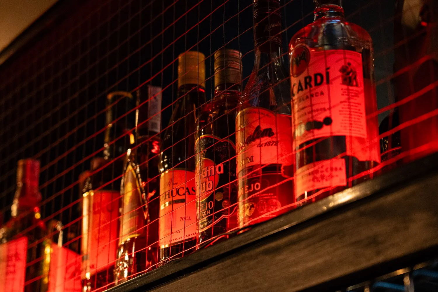 Botellas de alcohol, principalmente ron, en una estantería detrás de una reja en un bar oscuro, con iluminación rojiza.