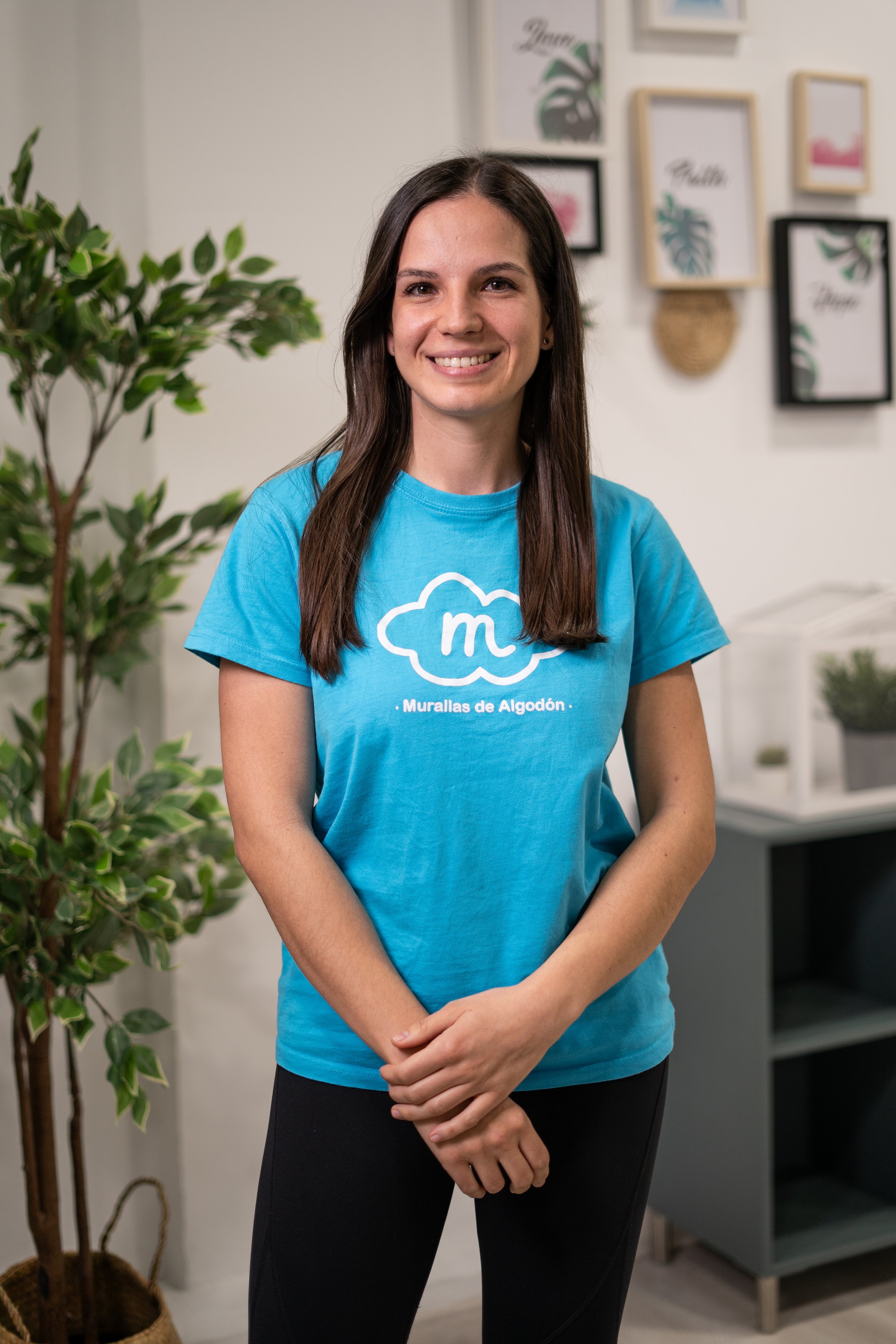 Mujer joven sonriendo, lleva camiseta azul con texto y graphic de nube, en un ambiente interior decorado con cuadros en la pared y planta en una maceta.