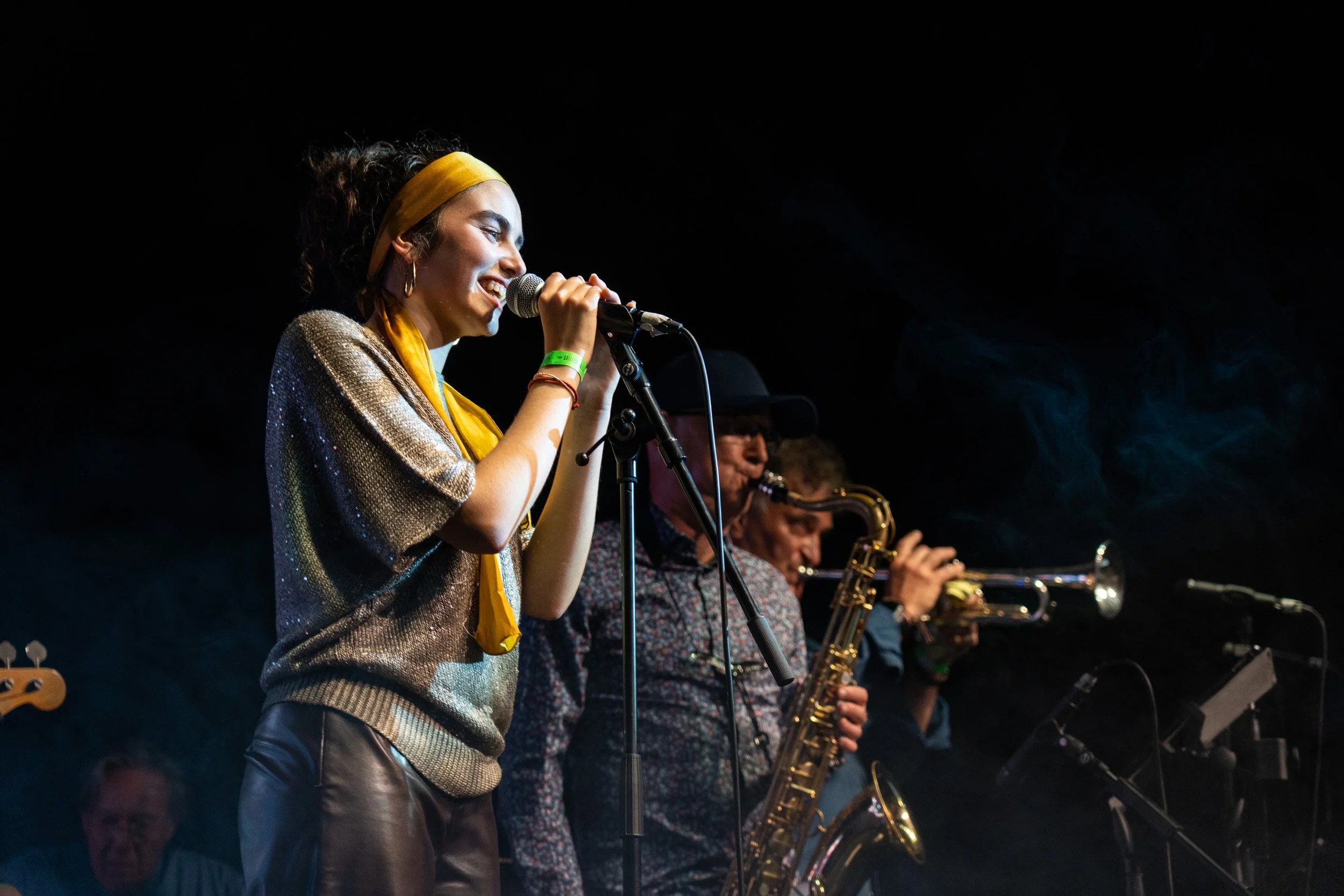 Mujer cantando con banda de músicos, en escenario oscuro, tocando instrumentos como saxófonos.