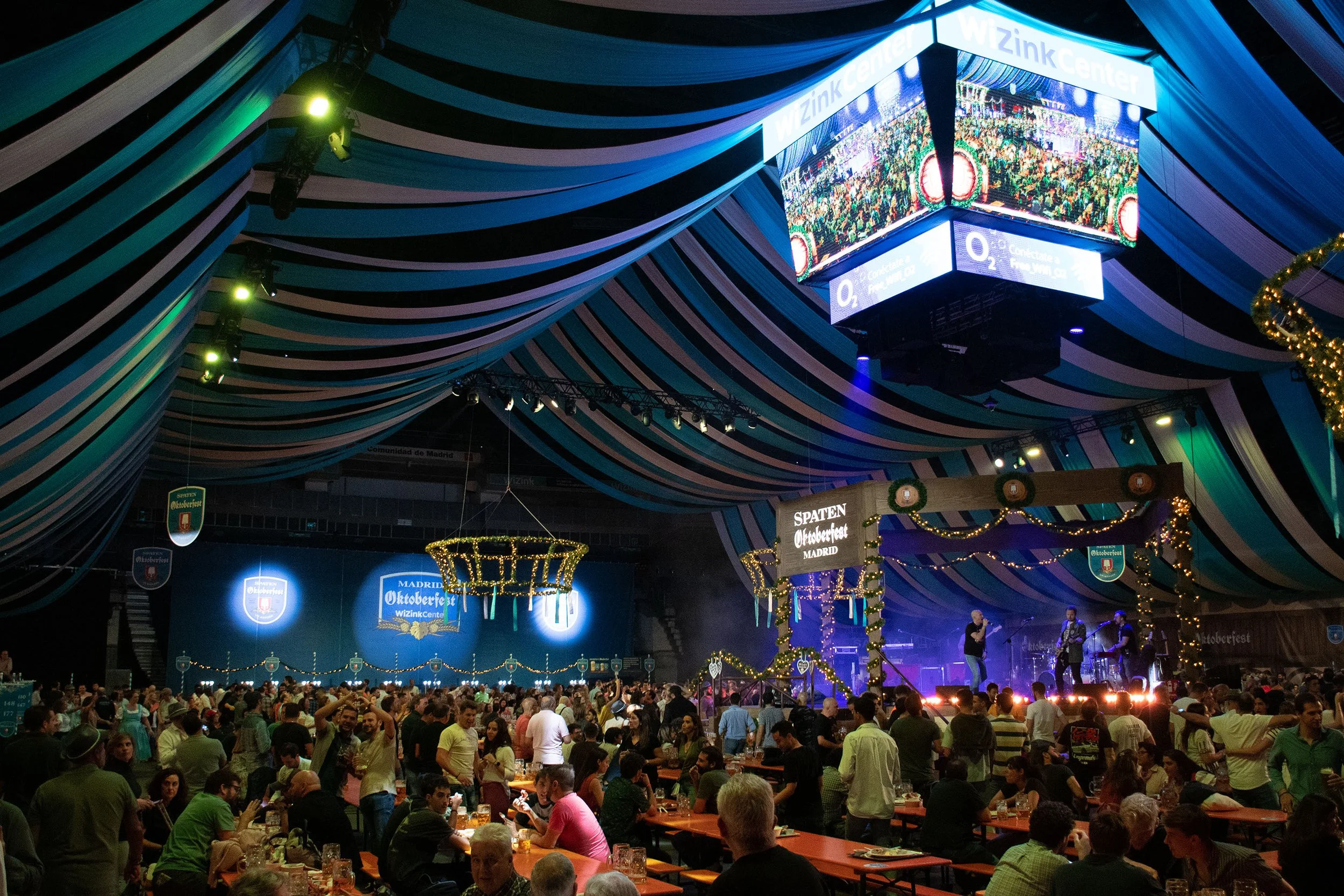 Interior de una tienda o evento con decoraciones tradicionales bávaras, con un gran número de personas, mesas de madera, banderas y un gran monitor con el texto 'WIZINK CENTER' y una imagen de una multitud. Ambiente festivo con iluminación tenue y decoraciones navideñas.