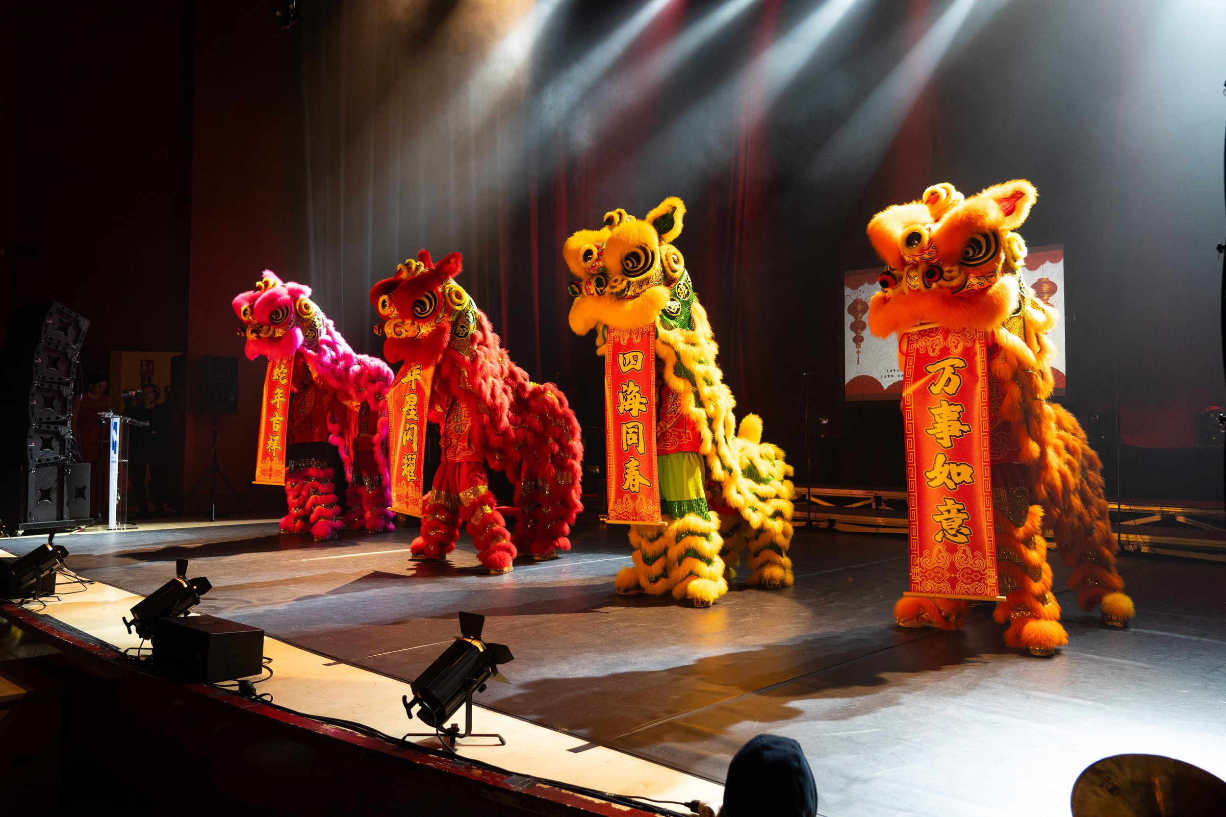 Presentación de bailarines con disfraces de leones tradicionales en un escenario con iluminación de luces doradas y fondo oscuro.