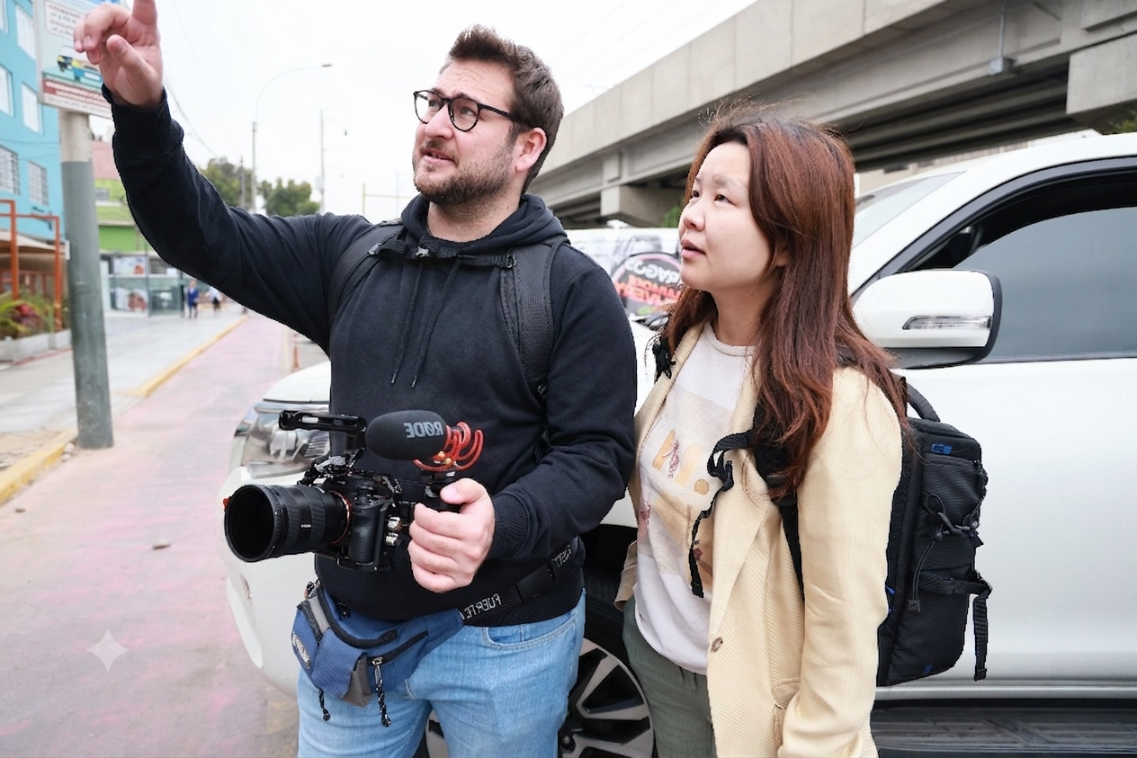 Dos personas, un hombre con gafas y una cámara en la mano, David Amar Foto, y una mujer asiática con mochila, están conversando y señalando algo en la calle, junto a un carro y en un día nublado.