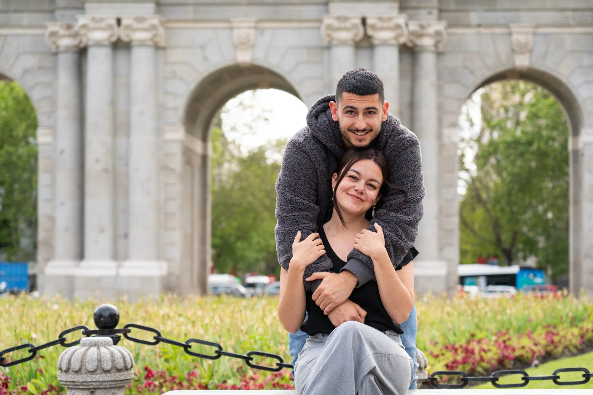 Una pareja joven en un parque, la mujer sentada y el hombre apoyado sobre ella, ambos sonriendo. Detrás hay un arco de piedra y áreas verdes con flores.