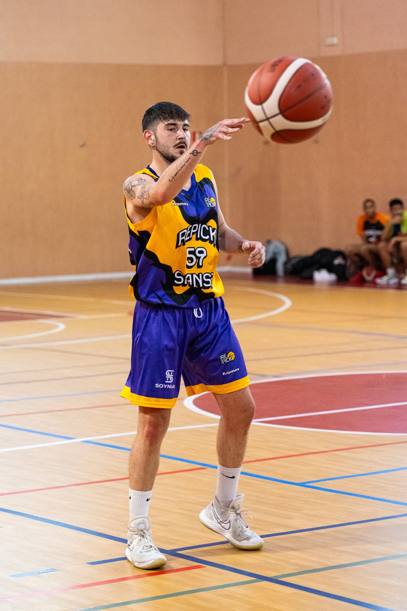 Joven jugador de baloncesto lanzando una pelota en una cancha cubierta, viste un uniforme amarillo y azul, con pantalones cortos, camiseta sin mangas y calzado deportivo blanco.