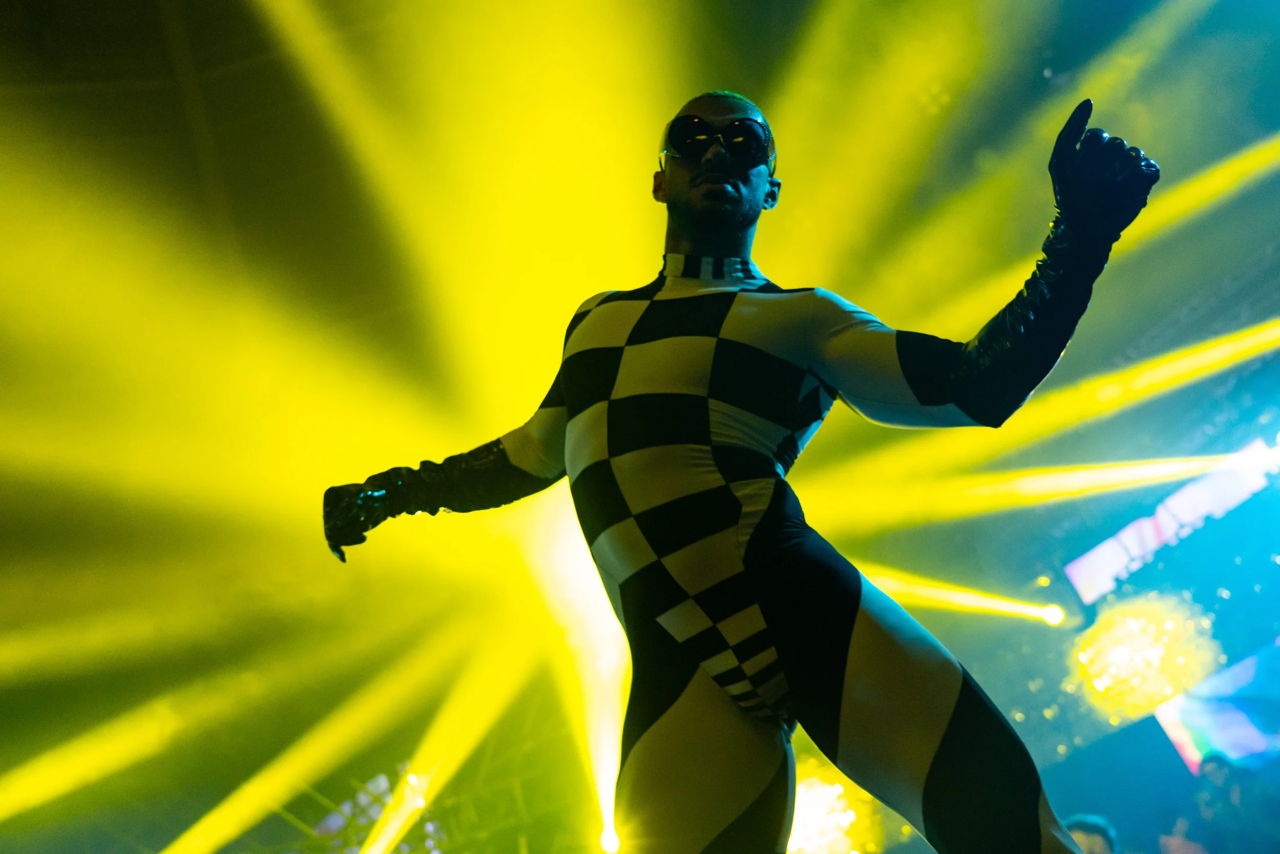 Una persona con traje de cuerpo completo de patrón de cuadros en blanco y negro, y guantes negros, en una escena con luces amarillas y verdes, y usa gafas de sol negras. Parece estar en un escenario o en una presentación artística o de moda.