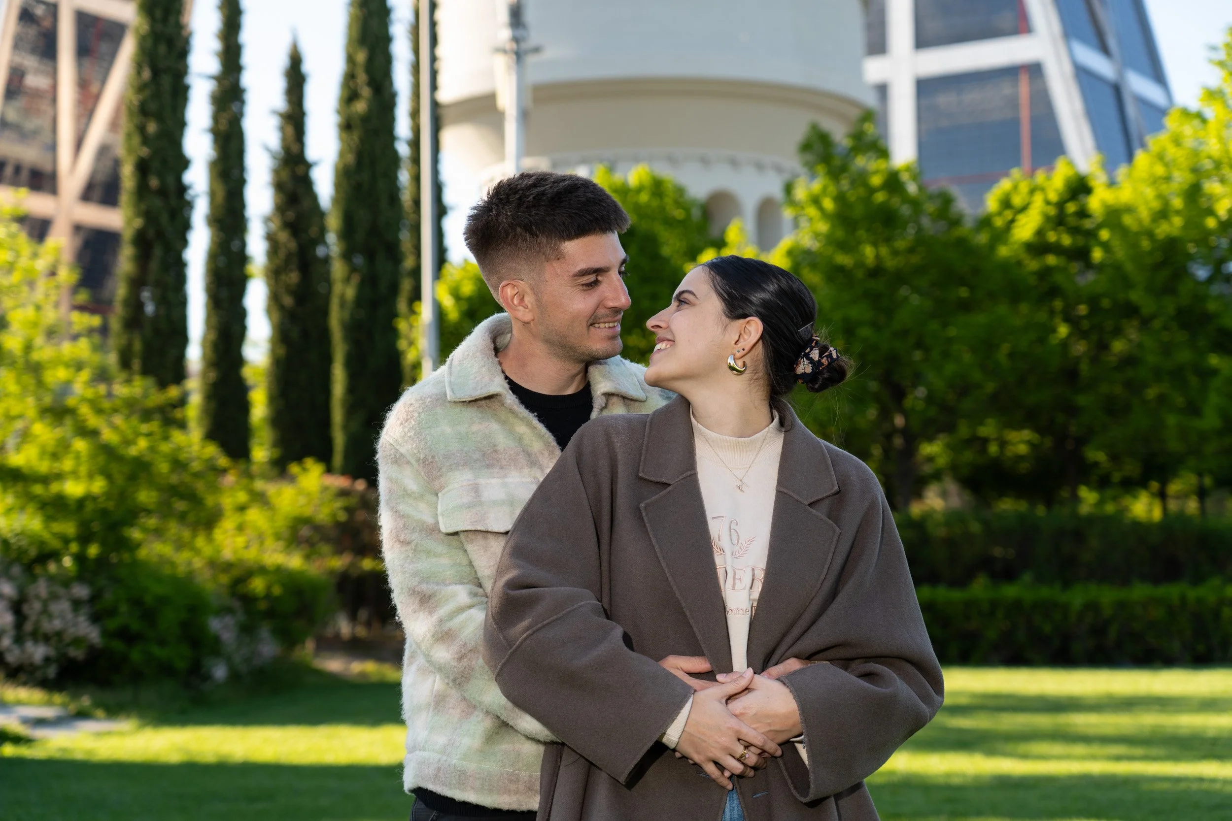 Una pareja en un parque, mirándose con cariño y sonriendo, rodeados de árboles y edificios modernos.