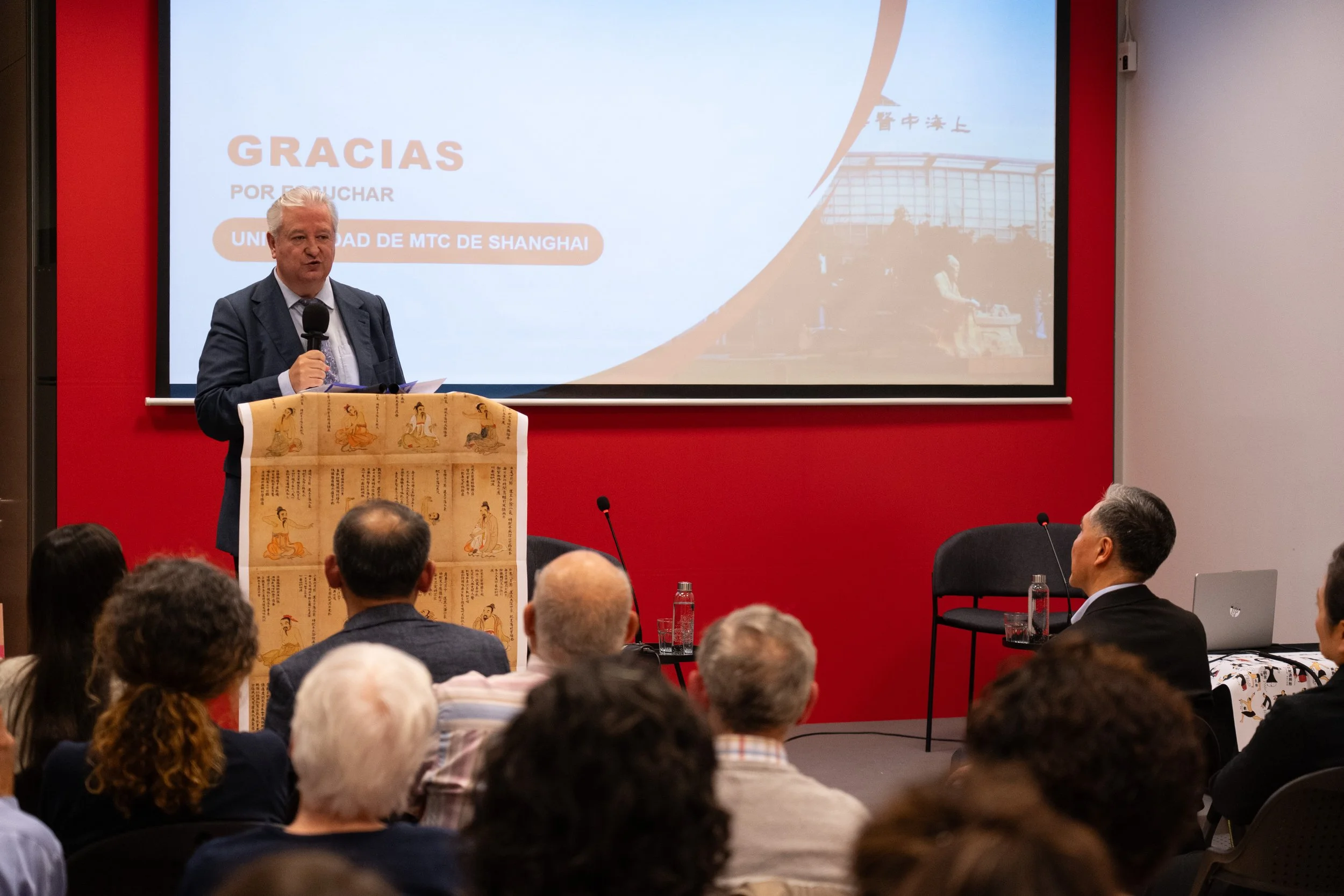 Hombre en traje, de cabello rubio, hablando en una conferencia frente a un micrófono y una pantalla que dice "Gracias por su atención Universidad de MTC de Shanghai". La audiencia está sentada y hay una pantalla con una imagen del edificio de la universidad en el fondo.