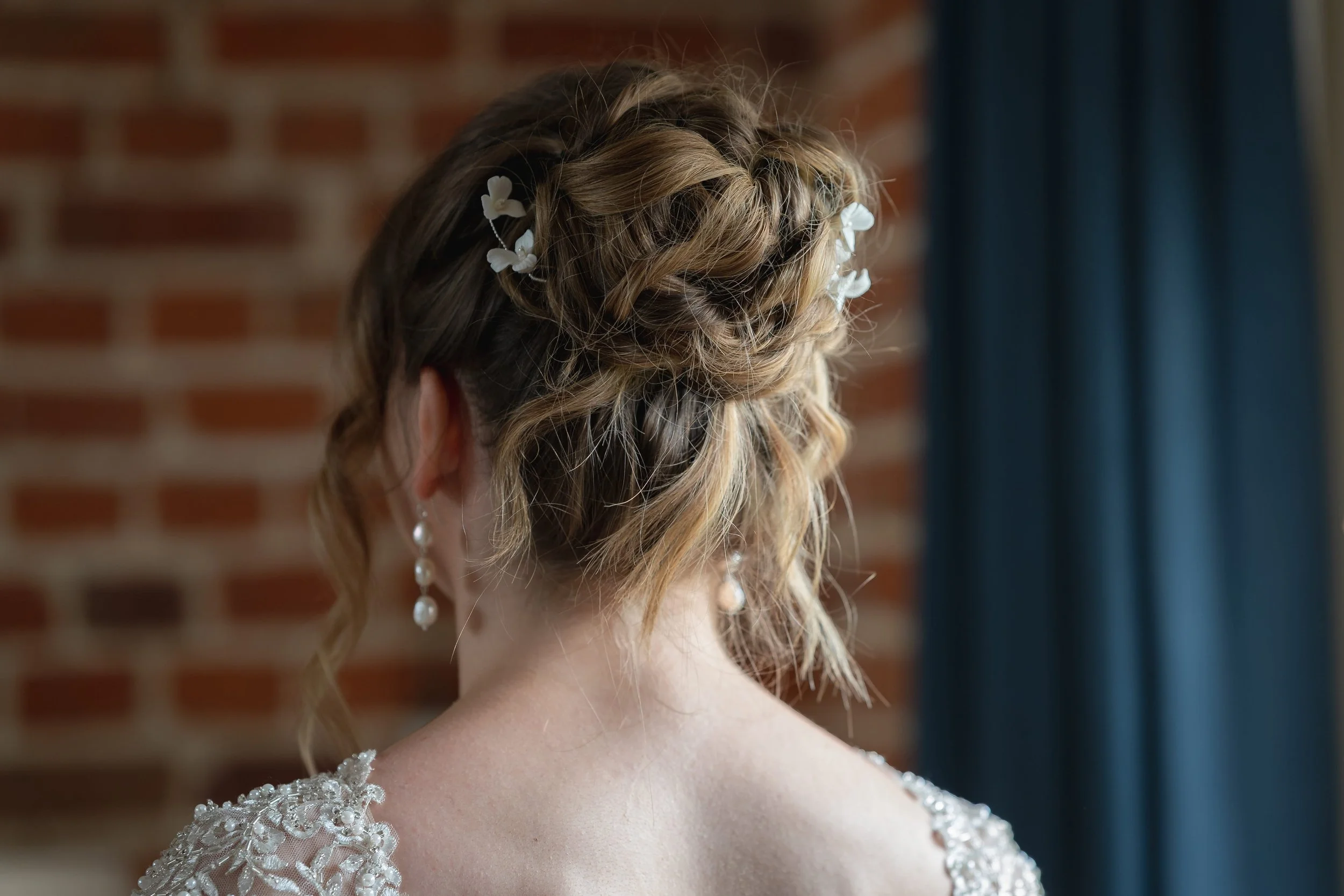 A bride facing away from the camera, wearing her hair in a textured high bun with white accessories and pearl earrings, ready for her wedding day at Channels Estate in Chelmsford, Essex.