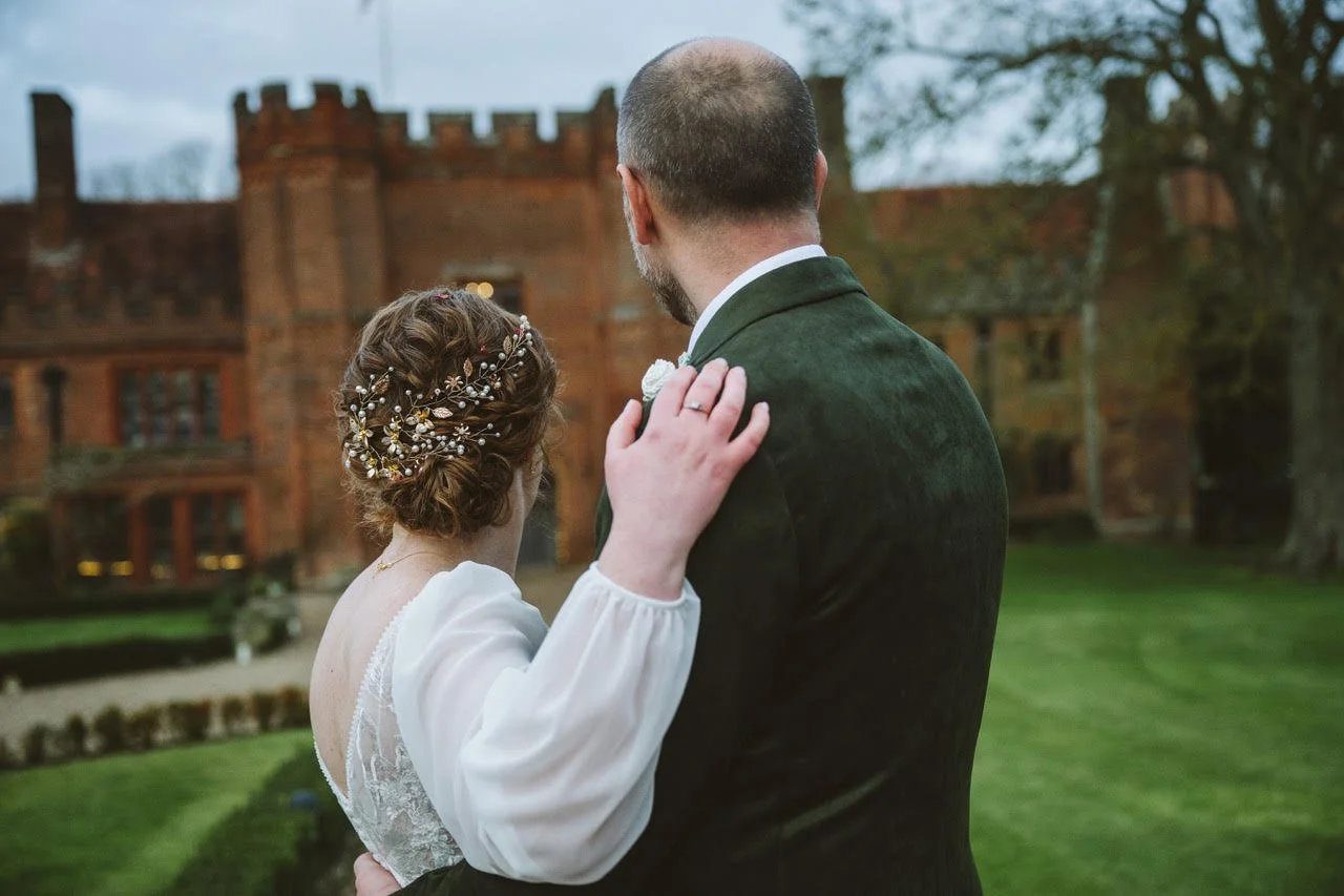 A back view of a bride and groom embracing, looking towards their wedding venue (Leez Priory in Chelmsford). The bride is wearing her hair in an updo using her natural curls, with a beaded hair accessory.
