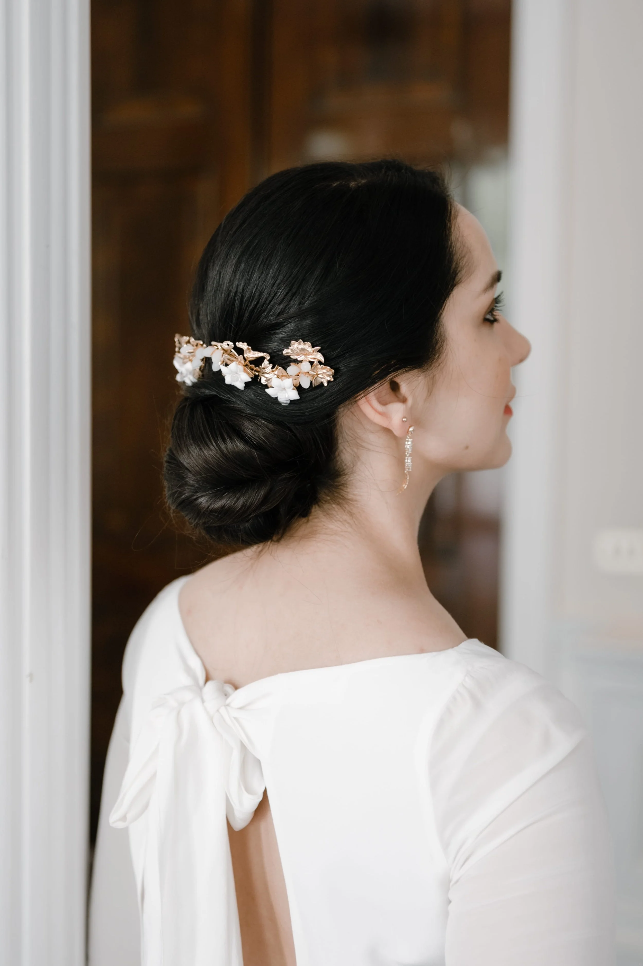 A photo of a bride taken from behind, wearing her dark hair in a clean low bridal bun with gold and white floral accessory, ready for her wedding day in London.