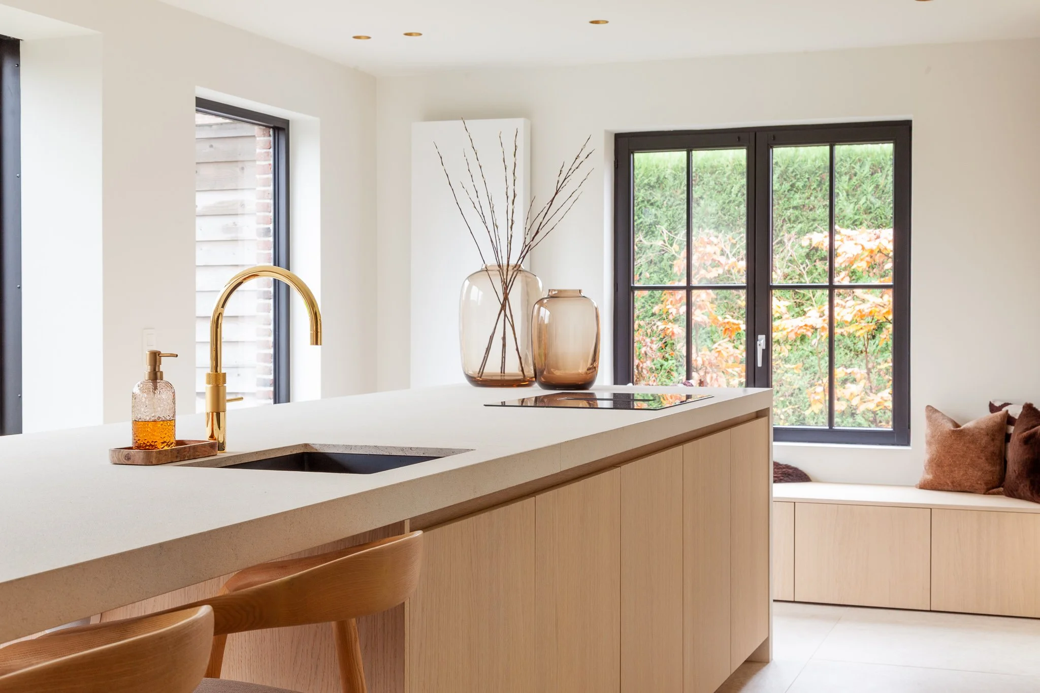 Moderne keuken met een witte keukenblad, gouden kraan, twee grote glazen vazen met takken, grote ramen met uitzicht op een tuin met herfstkleuren en comfortabele bankje met kussens.