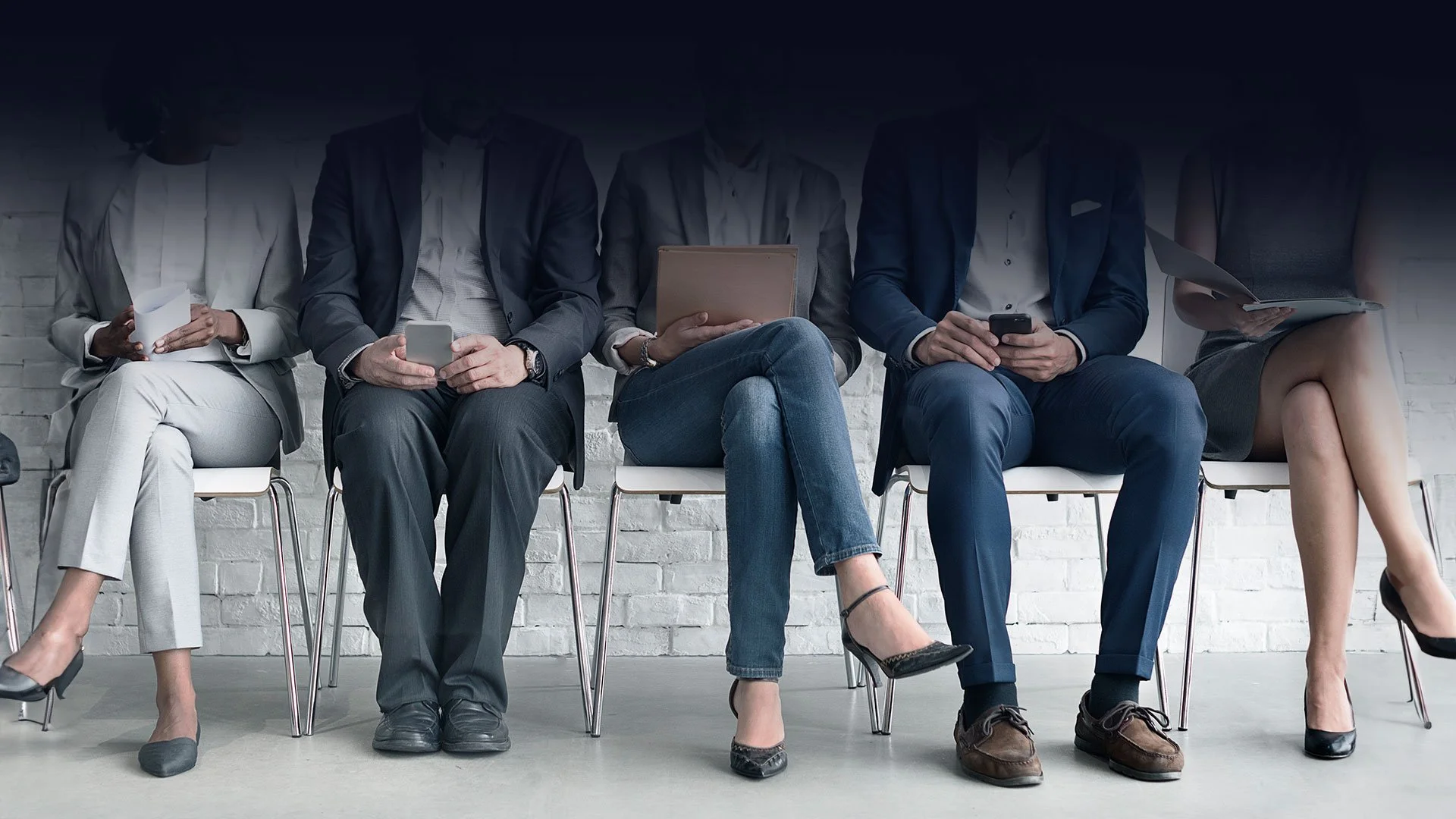 Five diverse people sitting in chairs against a white brick wall, dressed in business attire, engaging with their smartphones, tablets, or documents.