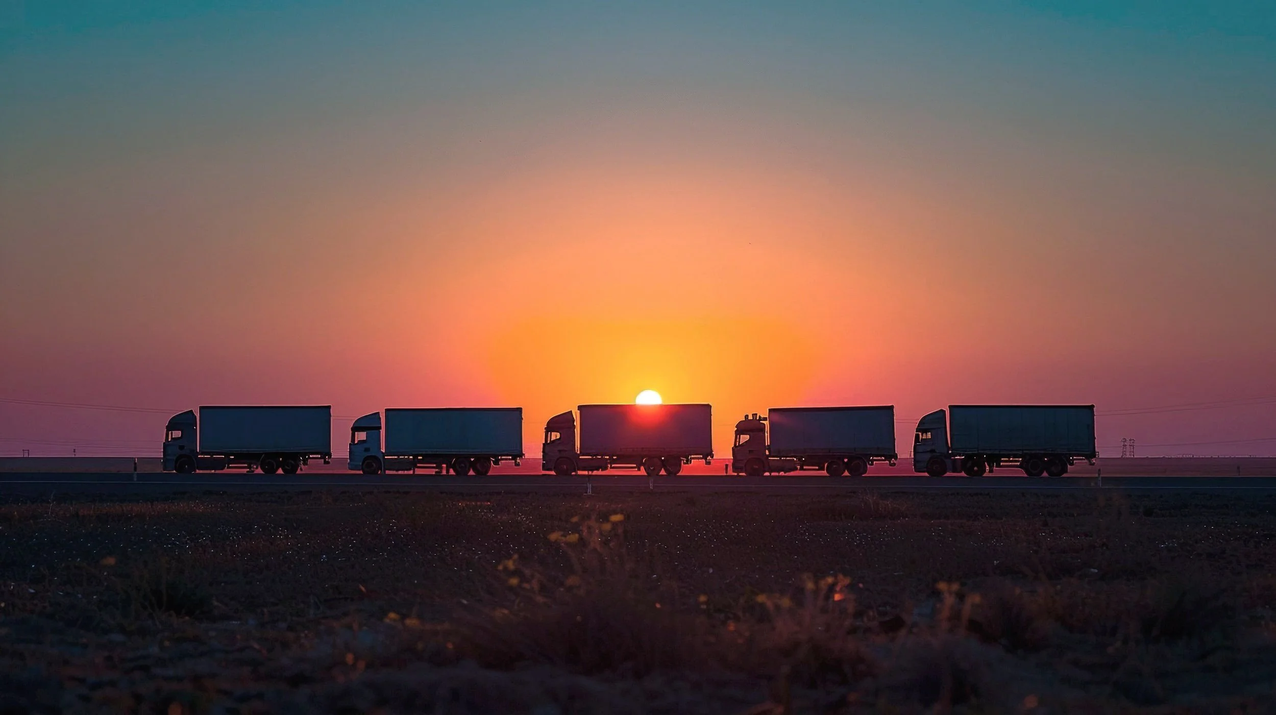 Five trucks driving on a highway at sunset, with a colorful sky in the background.