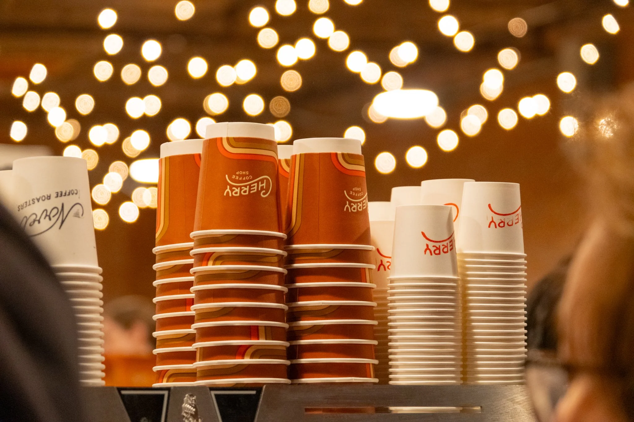 Stacked paper coffee cups with orange and white branding on a counter, with blurred warm string lights in the background.